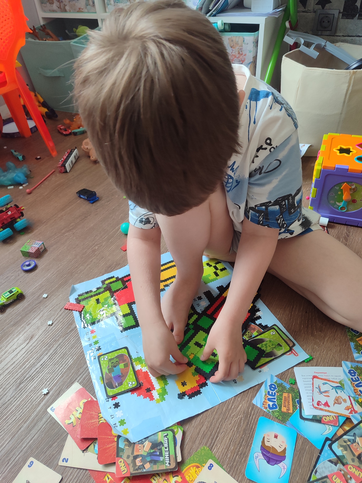 A child with short brown hair sits on a wooden floor, engaged in building a pixelated image with small plastic squares. The image is on a blue piece of paper marked with numbers. Surrounding the image are various playing cards, including UNO and Minecraft-themed cards. Toys like miniature vehicles and building blocks are scattered nearby. The indoor environment contains storage bins and other toys in the background. There is nothing to imply the photo was taken in Izhevsk, Russia.