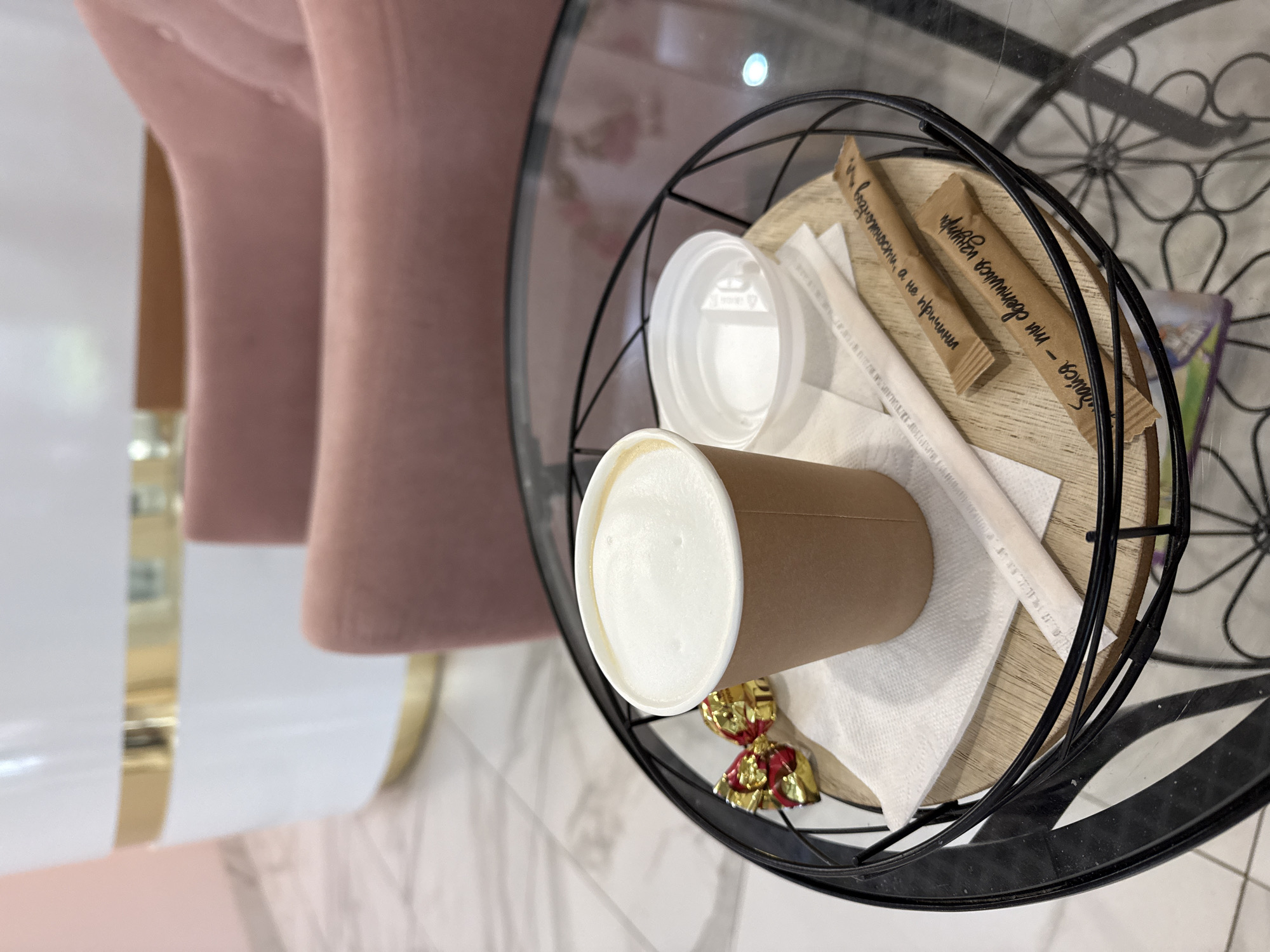 A close-up, eye-level shot captures a coffee cup and accompaniments on a round glass table with a black metal wire basket. The coffee cup, made of brown paper, is filled with a frothy beverage, likely coffee with milk. Next to it, a white plastic lid rests on a wooden coaster. Two individually wrapped sugar packets, one with Russian text that translates to "Smile - you are a gem," and a straw are also on the coaster. To the left of the coffee cup, two gold and red wrapped candies are placed on a white paper napkin. In the blurred background, a portion of a white and gold reception desk or counter is visible, along with the soft pink upholstery of two chairs. The floor is tiled with a marble-like pattern.