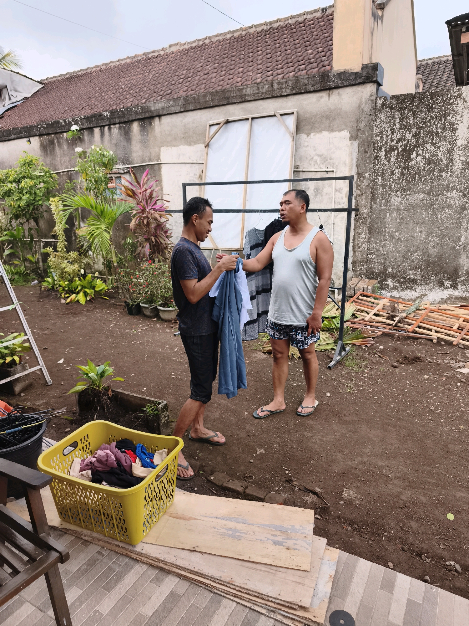 In Kaligondo, Indonesia, two adult males are engaged in sorting laundry outdoors. One man, wearing a dark t-shirt and shorts, holds a blue garment. The other man, in a light-colored tank top and patterned shorts, stands facing him, also holding a garment. They are positioned in a dirt yard with a clothesline rack behind them, upon which several articles of clothing are hung. In the foreground, a yellow laundry basket overflows with folded clothes. The background features a building with a tiled roof and lush green vegetation. The scene suggests a domestic chore being performed in a casual, outdoor setting.