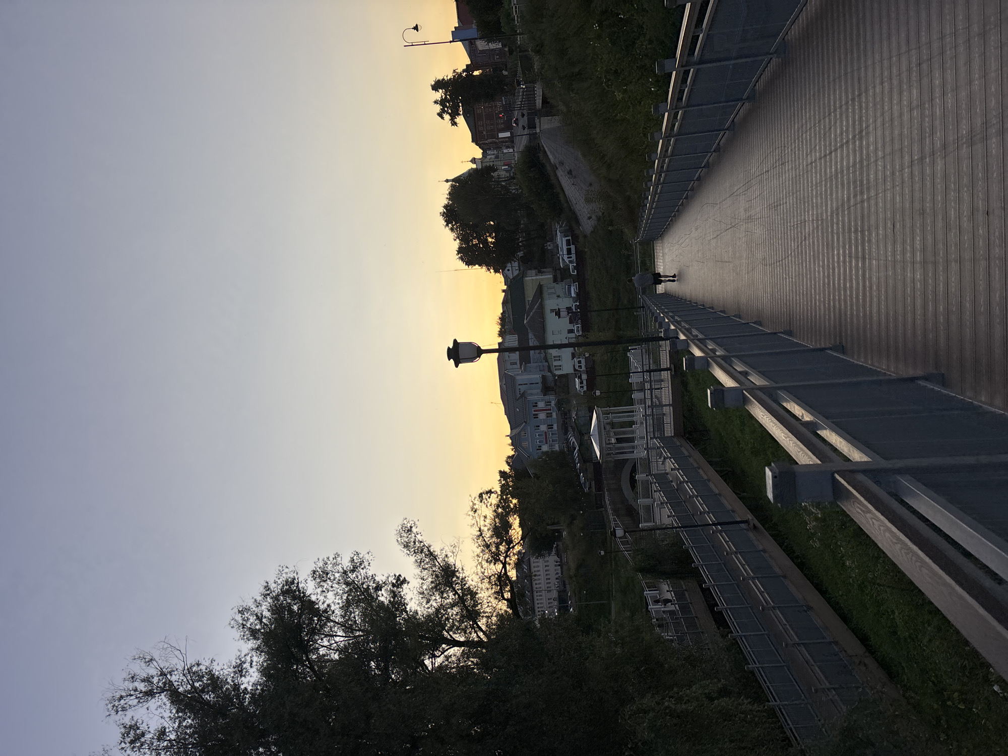 This is a vertical, eye-level, medium shot of a wooden pedestrian bridge in Klin, Russia, at dusk. The sky is a pale gradient from light blue at the top to a warm yellow-orange near the horizon, indicating sunset. A large tree with dark green foliage is visible on the left side of the frame. The bridge, made of horizontal wooden planks and with metal railings, extends into the distance and curves slightly to the right. A lone figure, seen from behind, walks away from the camera on the bridge. Below the bridge, there is a green, grassy area with some vegetation and another section of a walkway with metal railings. In the background, across a river or canal, are buildings of various sizes and architectural styles, some with red roofs, and more trees. A white gazebo-like structure with columns is also visible in the middle ground. The lighting is dim, consistent with the time of day.