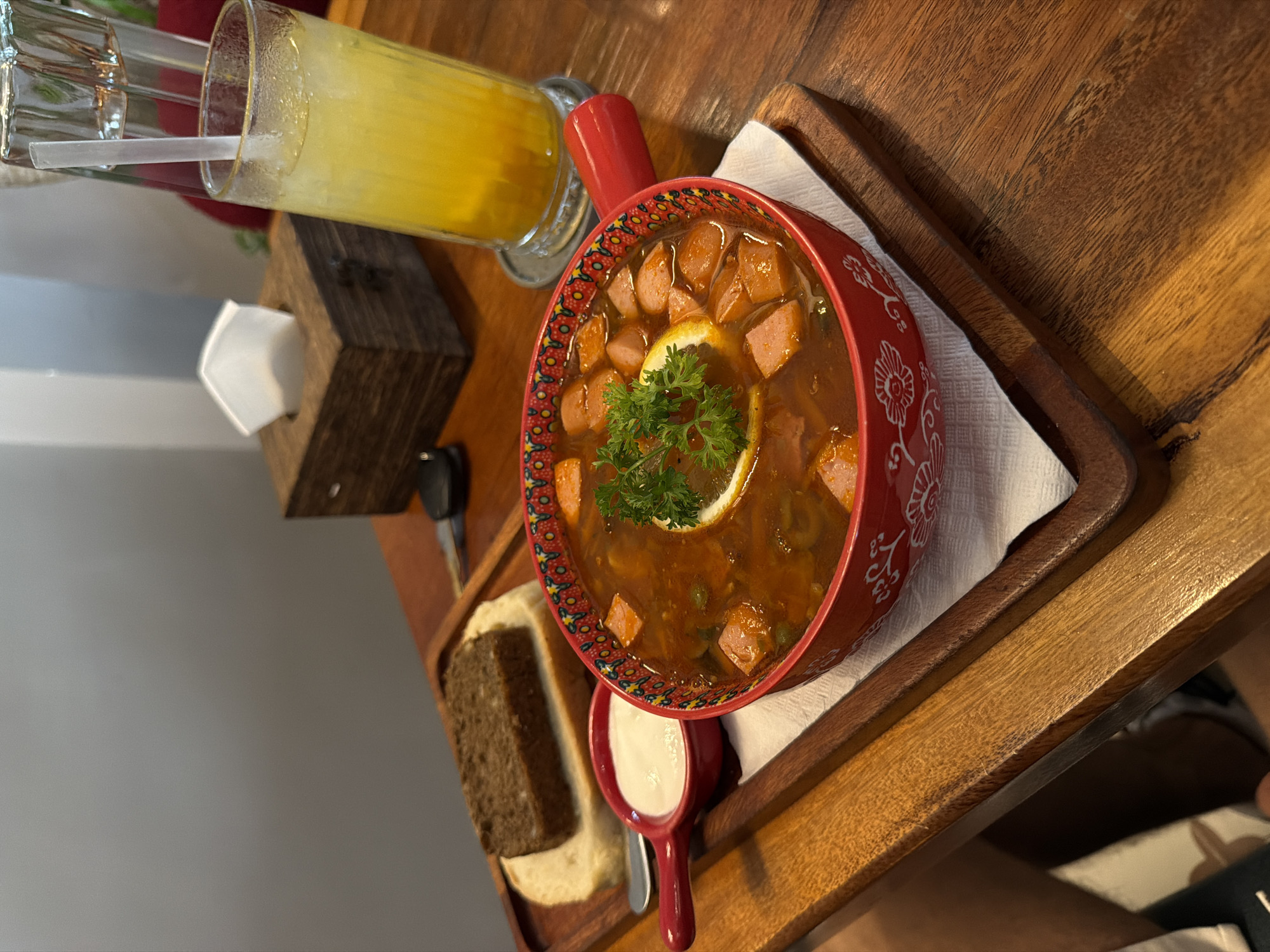 An overhead shot shows a wooden tray on a wooden table. On the tray are a bowl of soup, a small dish of white sauce, and a slice of bread topped with a darker slice of bread. The soup bowl is red with white floral patterns. The soup is a reddish-brown broth with diced pieces of sausage, peas, and a slice of lemon and parsley on top. The small dish contains a white, creamy sauce. The bread is a light-colored slice topped with a dark slice. To the right of the tray is a glass of yellow liquid with ice and a straw. Behind the glass, a patterned pitcher can be seen. Further back, a wooden tissue box sits on the table. A key is partially visible on the table near the tissue box. The table appears to be in an indoor setting, possibly a restaurant in Da Nang, Vietnam, given the context.