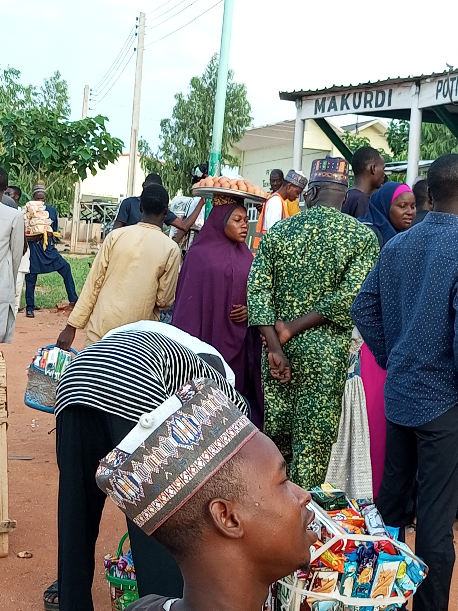 In Gombe, Nigeria, a group of people are gathered outdoors, with a sign overhead that reads "MAKURDI POTH". Several individuals are dressed in traditional attire, including a person wearing a vibrant green and yellow patterned garment and another in a purple hijab. A woman is balancing a tray of eggs on her head, indicating a market or roadside sale. Bags of goods, possibly snacks or food items, are visible, held by a person in a striped shirt and a basket in the foreground. The ground is covered in reddish-brown dirt, and there are trees and utility poles in the background, suggesting an urban or semi-urban environment. The scene appears to be a bustling commercial area with active transactions taking place.
