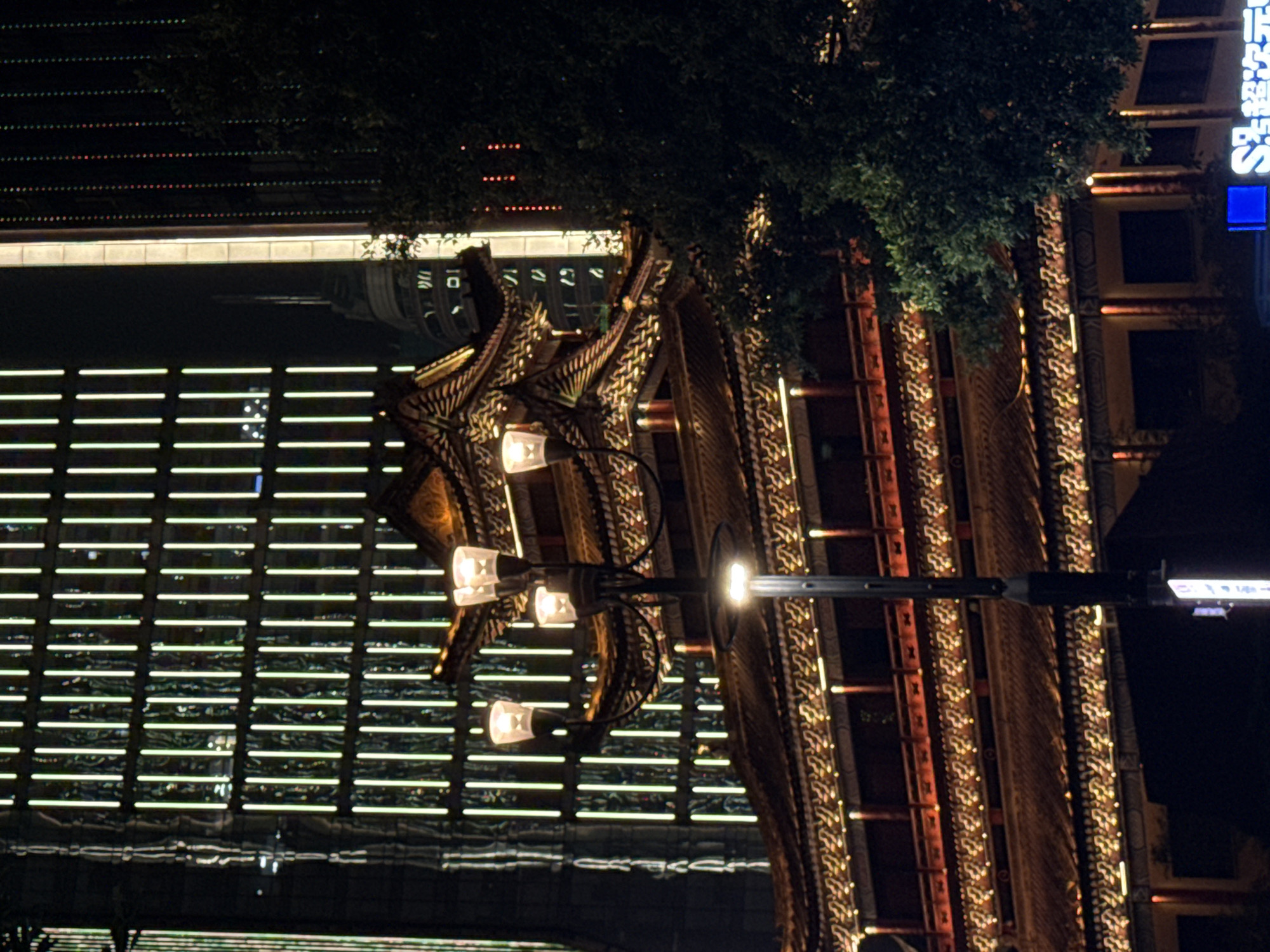 In Yuzhong District, China, at night, a modern skyscraper with vertical white lights stands behind a traditional Chinese building with a tiered, ornate roof. A streetlamp with multiple glowing bulbs is positioned in the foreground, partially obscuring the traditional structure. The sky is dark, and the overall scene is illuminated by artificial lights from the buildings and the streetlamp, creating a contrast between modern and traditional architecture.