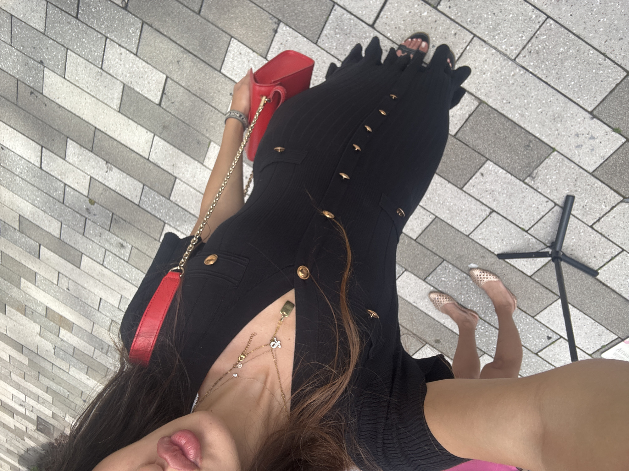 A person is standing outdoors on a paved surface composed of rectangular grey and white tiles. They are wearing a black, ribbed, form-fitting dress with gold buttons. A red handbag with a gold chain strap is slung over their shoulder. The person is also wearing jewelry, including a gold necklace with multiple charms and a bracelet on their wrist. Their feet are adorned with rose gold flat shoes and black sandals. The background consists of the tiled ground, with the person's legs and arms partially visible. The image captures a section of the person's face, showing their lips. The scene appears to be a public area, potentially in Aventura, Florida, United States of America, given the context.