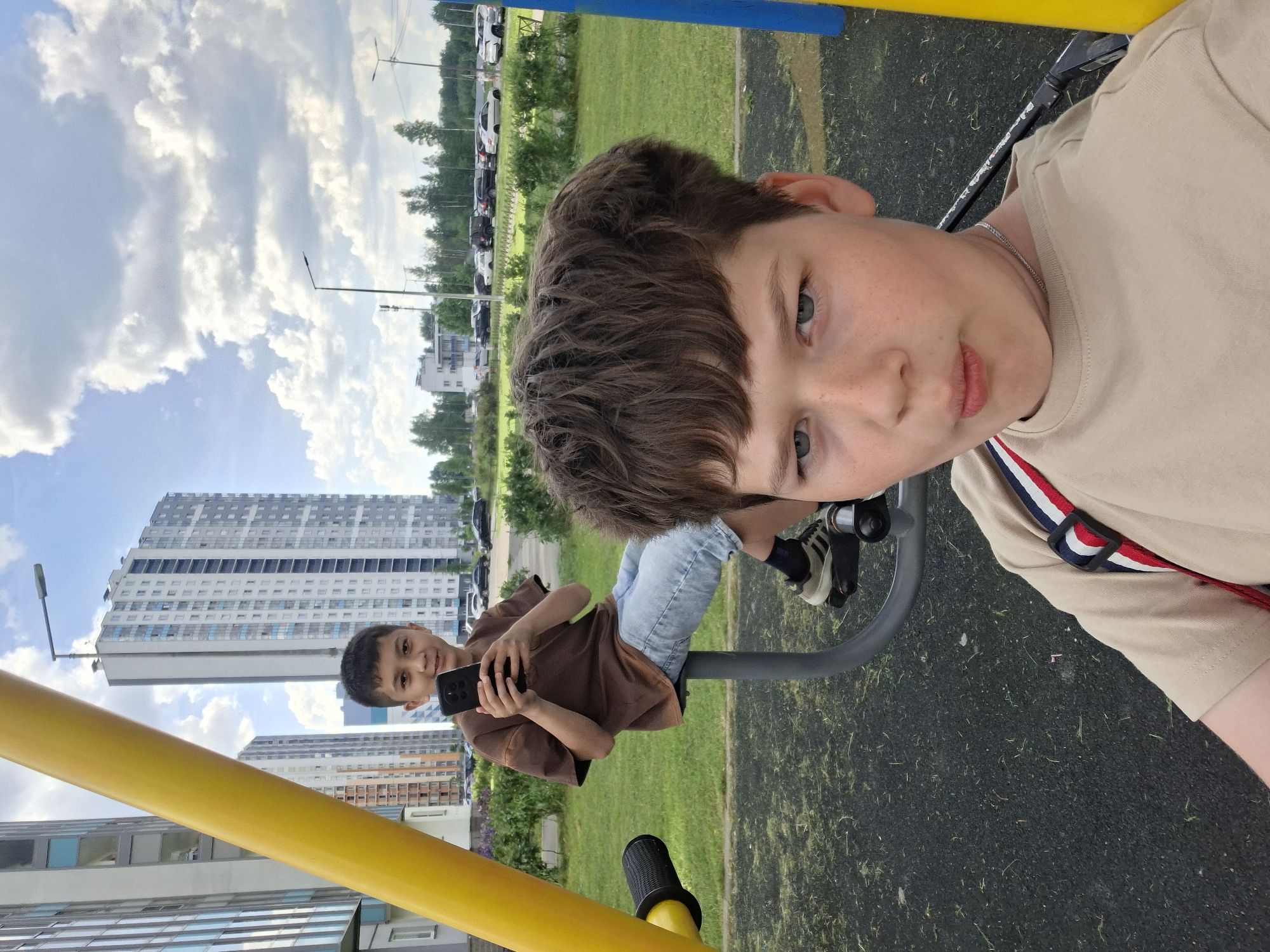 The image, rotated 90 degrees counter-clockwise, captures two boys at an outdoor playground in Saint Petersburg, Russia, during daytime under a partly cloudy sky.
In the immediate foreground, a boy with dark brown, wavy hair and blue-green eyes looks directly at the camera. He wears a light tan t-shirt, a silver chain necklace, and has light freckles across his nose and cheeks. A red, white, and blue striped strap is visible on his right shoulder, possibly from a bag or part of his clothing. He appears to be posing for a selfie or being photographed.
Further back, another boy is seated on gray playground equipment, holding a black smartphone. He wears a brown t-shirt and blue jeans, and has dark hair. He is looking towards the foreground boy with a slight smile.
The setting is a modern urban playground, featuring sections of green grass and a dark, rubberized safety surface. Bright yellow and blue metal bars of playground structures are visible. In the background, tall residential buildings with numerous windows rise against the sky, characteristic of contemporary city architecture. Trees and parked cars are also visible in the distance, suggesting a suburban or urban edge environment. No legible text is discernible within the image.