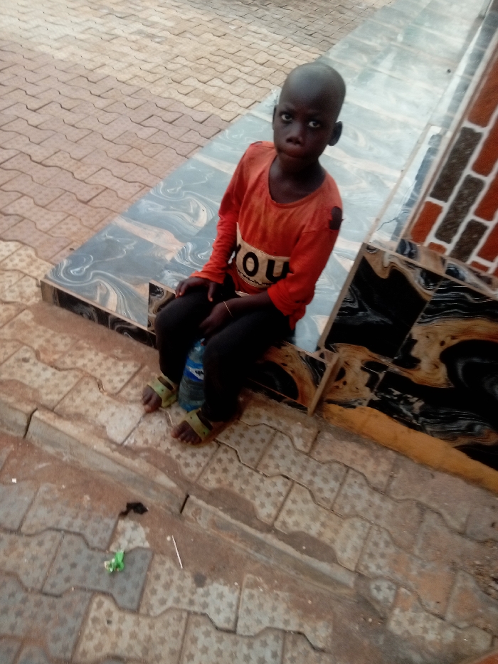 A young child is seated outdoors, looking directly at the viewer with a serious expression. The child has dark skin, closely cropped or shaved dark hair, and dark eyes. They are wearing an orange long-sleeved shirt that appears somewhat worn, with a white and black graphic on the front that reads "YOU." The child also wears dark pants and light green, open-toed sandals. A clear plastic water bottle with a blue cap is held between their legs.
The child is sitting on a low, tiled ledge or step. The tiles on this ledge and an adjacent structure are dark, featuring a swirling pattern of black, gray, and possibly white or beige. Behind the child and to their right, another part of this structure rises, topped with reddish-brown, brick-like textured tiles.
The ground in front of the child and extending into the foreground is paved with interlocking concrete pavers. These pavers are light brown or beige and feature a repetitive star-shaped pattern. The ground appears somewhat dusty or covered with light dirt in areas. There is a small piece of green debris and a thin white stick or straw visible on the paving stones in the bottom left corner of the image. The overall lighting suggests it is daytime.