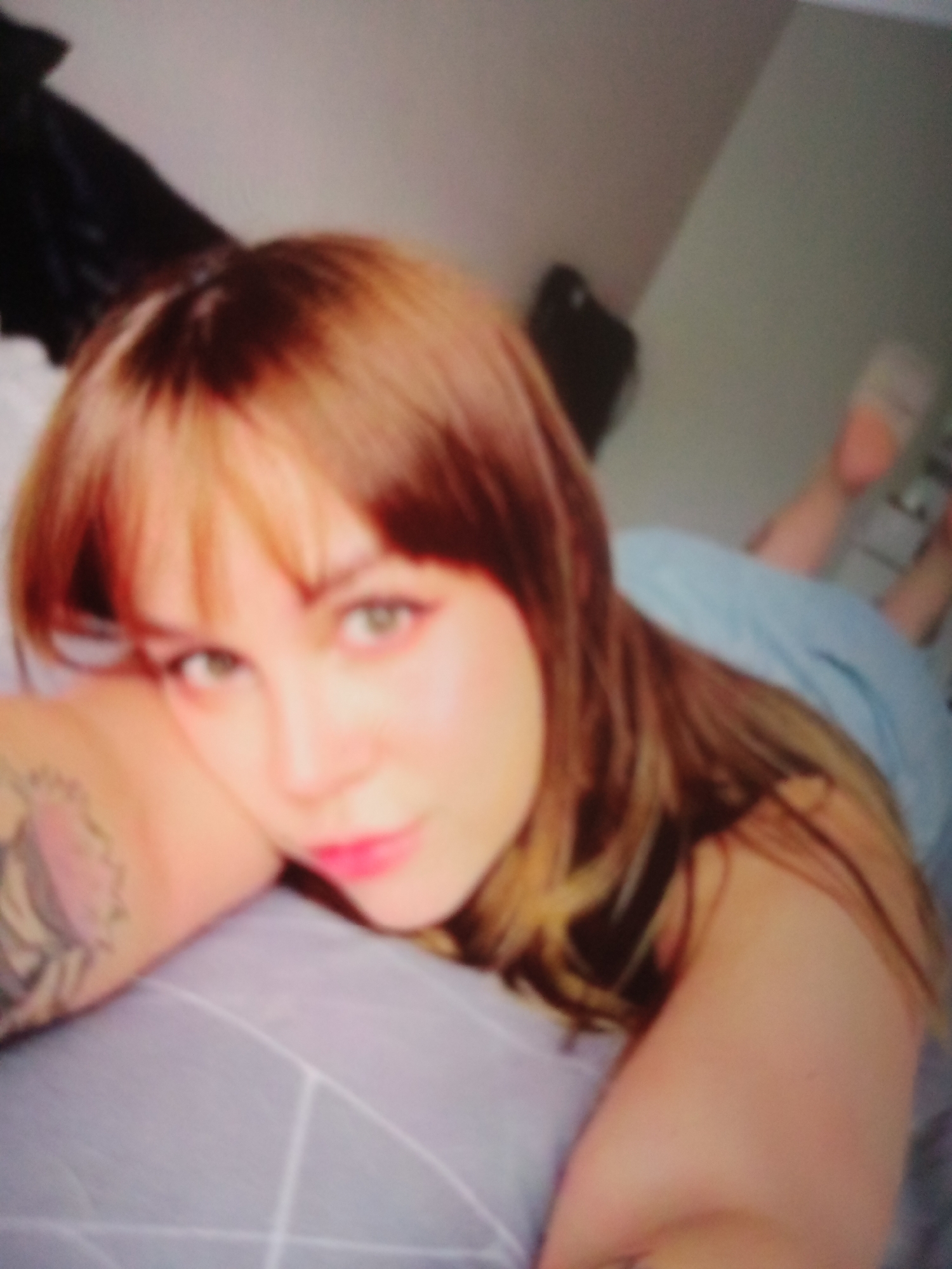 The provided image is a blurry, close-up, low-angle shot of a young woman lying down. She has light brown hair with bangs and is looking towards the camera with her head resting on her arm. Her eyes are green, and she is wearing bright pink lipstick. A tattoo is visible on her left arm. She is wearing a light blue garment, and the background appears to be a bed with a grey patterned sheet. There are no discernible landmarks or context that would allow for identification of the location in Jalingo, Nigeria.