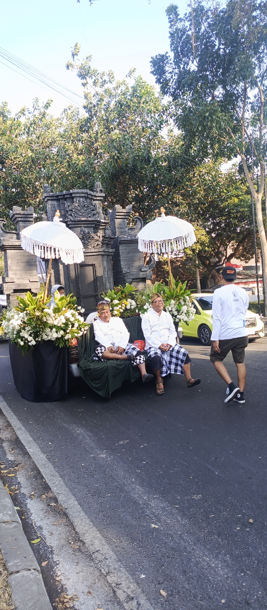 Two men in traditional Balinese attire, including headwear and checkered sarongs, are seated on a decorative float adorned with white flowers and umbrellas. They are positioned behind a large black structure covered in foliage. A man in a white long-sleeved shirt and dark shorts, wearing a black hat, stands facing away from the float, appearing to observe something to the right of the frame. In the background, a yellow car is visible, and behind it, a stone structure resembling a temple gate is present. Trees line the background, and the scene takes place on a paved road with a white line marking its edge. The presence of a similar float in a parade setting, combined with the architectural style of the gate, suggests this scene likely occurred in Malang, Indonesia, possibly during a cultural event or religious procession.