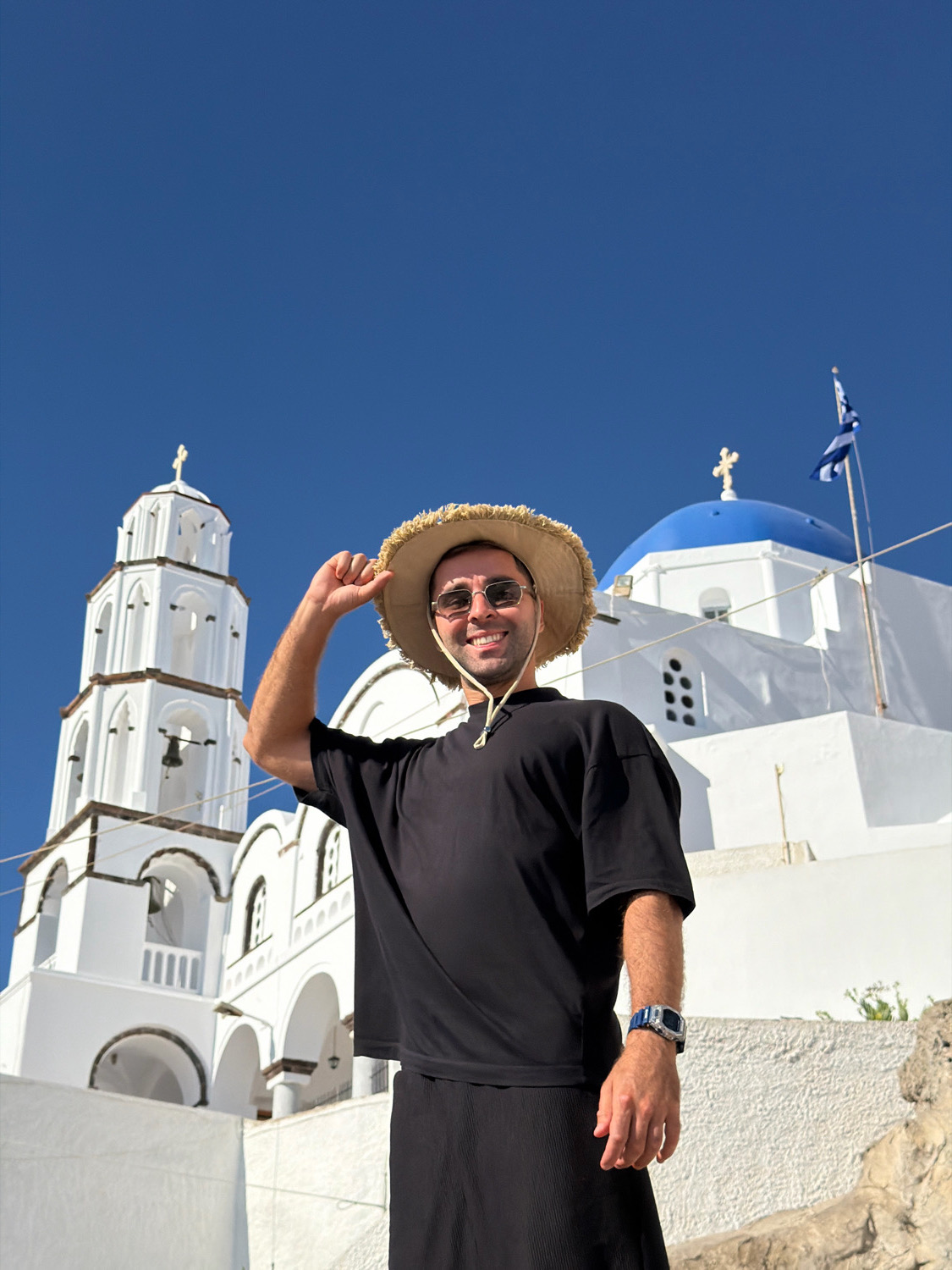 A man with a straw hat and sunglasses stands in front of a white church with blue domes in Thira, Greece. He is wearing a black t-shirt and black pants. The man is smiling and holding his hat. The church has a tall bell tower with bells and arches. The sky is clear and blue.