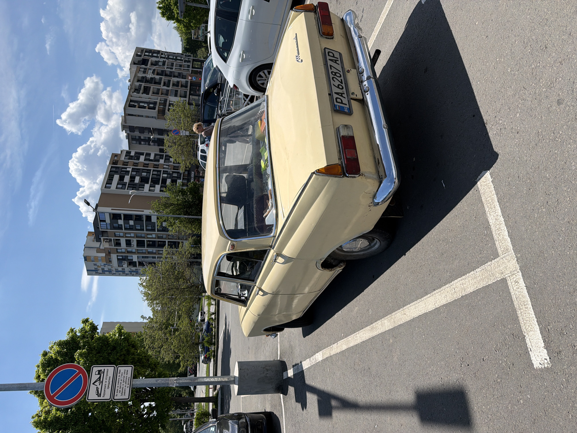The image depicts a beige Moskvich car parked in a parking lot in front of a modern building in Burgas, Bulgaria. The car's license plate reads "PA 6287 AP". Next to the parking spot is a sign indicating parking restrictions, likely with details regarding enforcement and time limits for customers of Lidl. Other cars are parked nearby, and the sky is blue with some clouds. The photo seems to have been taken during the daytime.
