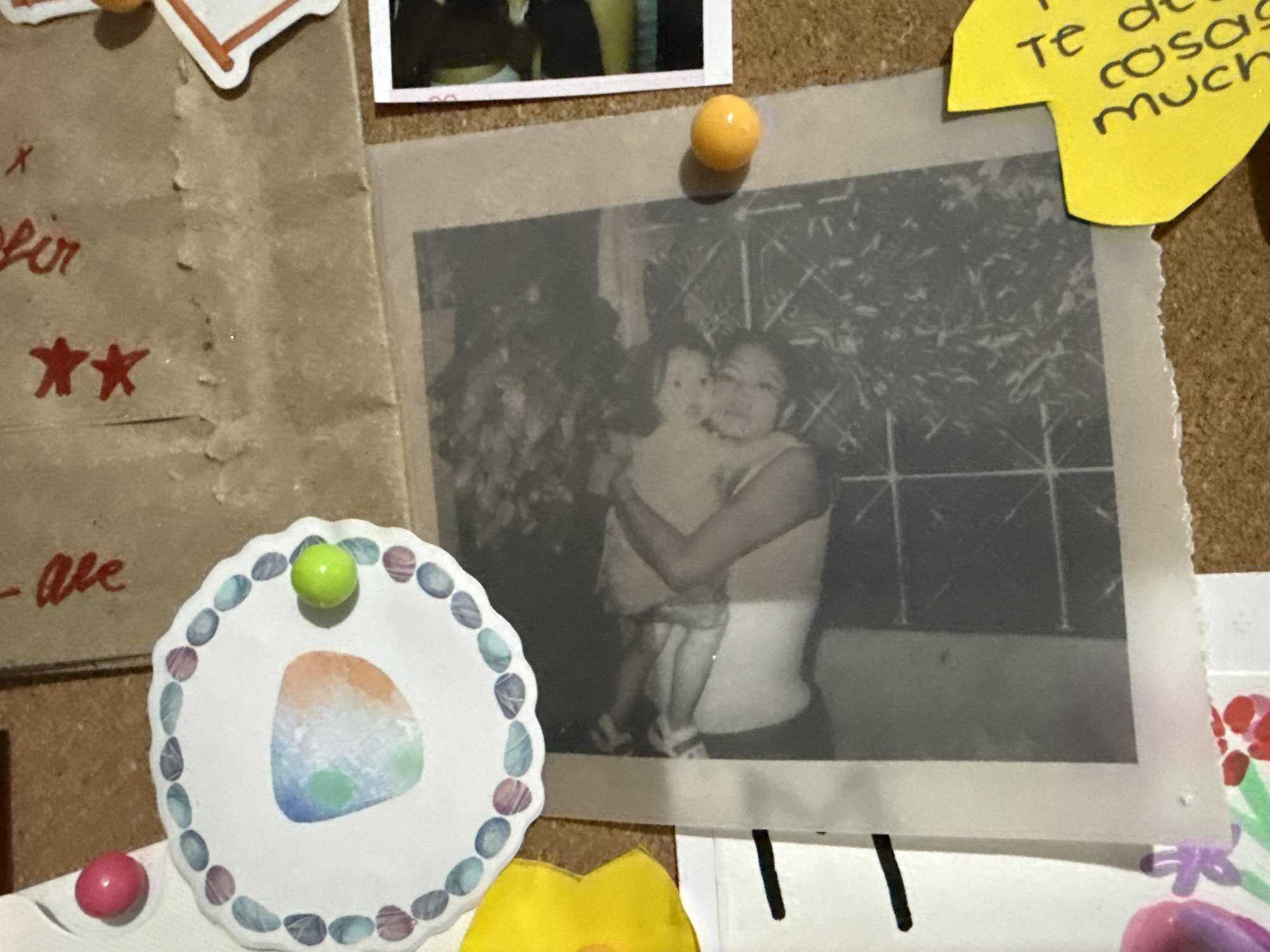 The image shows a portion of a bulletin board covered with various items. A black and white photograph depicts a woman holding a child, likely in a domestic setting, as suggested by the window and interior visible in the background. The photograph is attached to the board with a yellow pushpin. Adjacent to the photograph, a circular sticker with a colorful, abstract design is secured with a green pushpin. A portion of a brown paper bag is visible with red writing including the word "Ale" and two red stars. Other elements on the board include parts of a yellow paper cutout with black writing, a small snapshot, a sticker with a serrated edge, and several pushpins in various colors. The background material of the board appears to be cork. There is no contextual evidence in the image to suggest this is a photograph of Puebla, Mexico.
