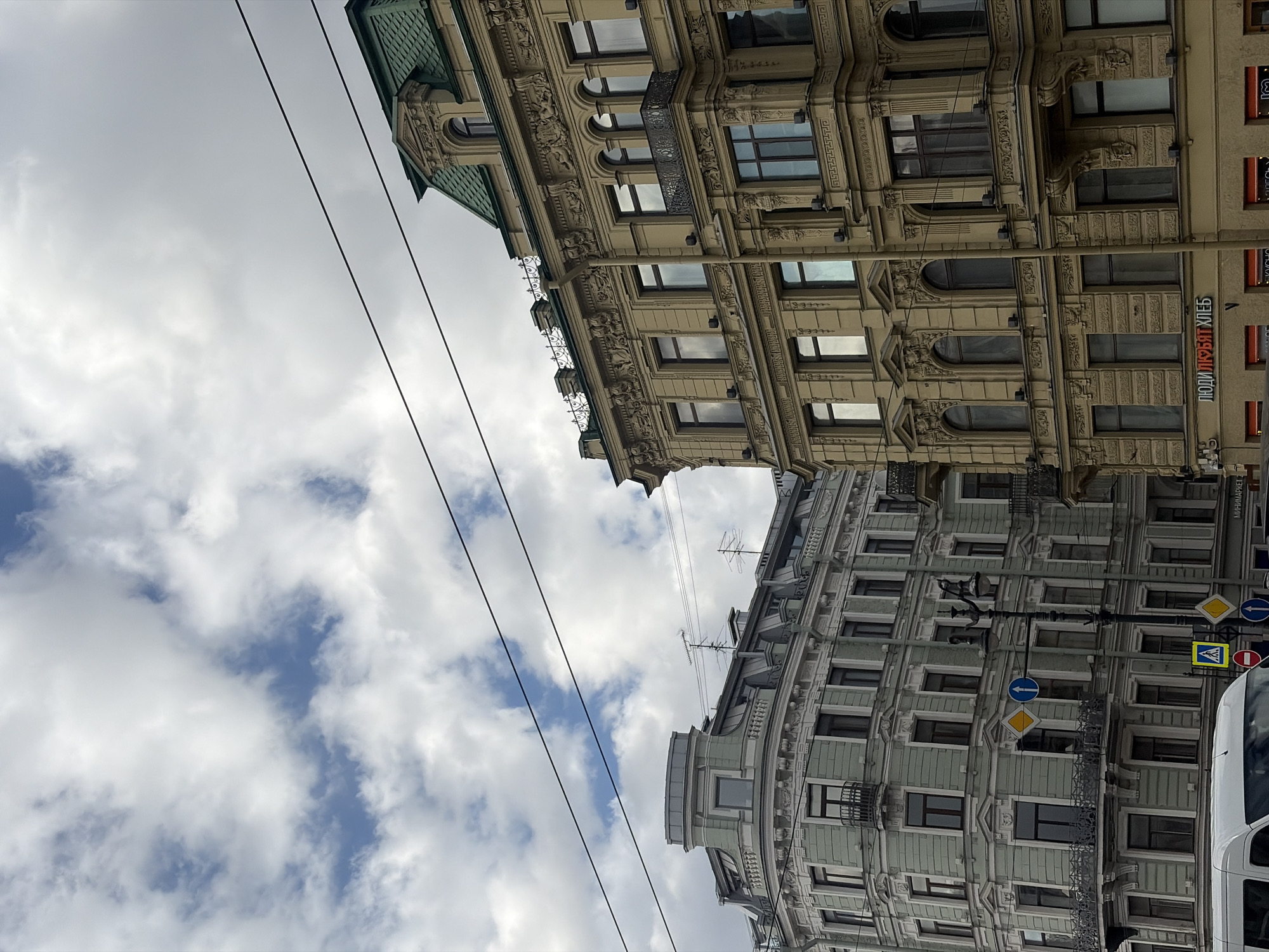 The image shows two buildings in Saint Petersburg, Russia, viewed from a low angle pointing upwards. The buildings are old and ornate, with detailed facades, many windows, and decorative elements. The building on the top is beige, and the building on the bottom is green. Power lines run diagonally across the image. The sky is filled with fluffy white clouds. Road signs and the side of a white vehicle are visible on the right. A sign with Cyrillic letters is also visible on the right building.