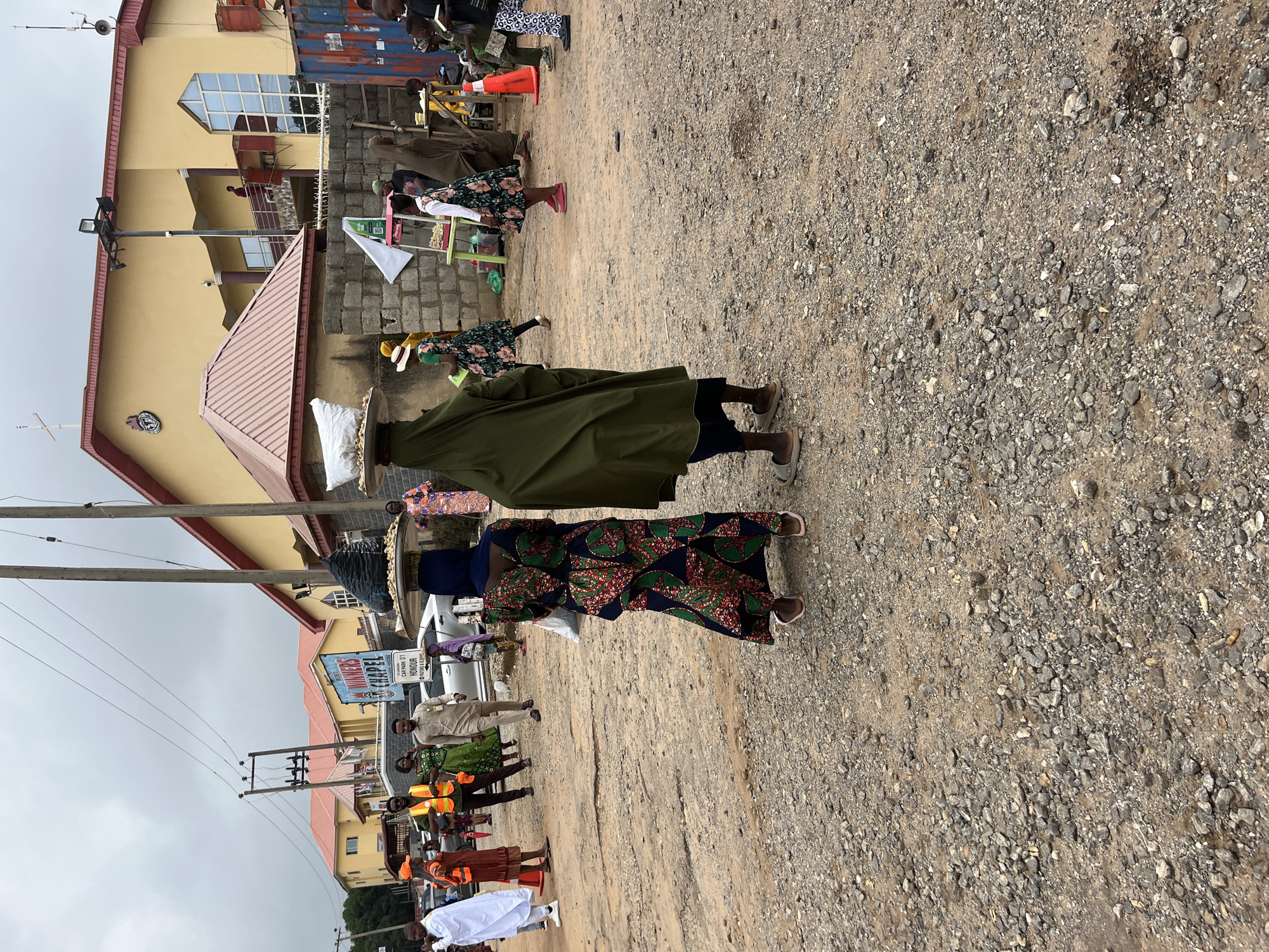 The image depicts an outdoor scene in a busy street or market area in Abuja, Nigeria. In the foreground, two women are walking away from the camera, each balancing a large tray of goods on their heads. The woman on the left wears a vibrant, multi-colored dress and a dark blue headscarf, while the woman on the right is draped in an olive green garment and a dark blue head covering. Their feet are visible, with sandals on the left and what appear to be open-toed shoes on the right.
The environment is a rough, gravelly ground, indicative of a less-developed area within the city. In the background, there are several buildings, including a prominent yellow building with a red roof, which appears to be a church, as evidenced by a cross on its upper facade and signage that reads "WINNERS CHAPEL." The building has modern architectural elements like large windows and balconies.
Several other people are present in the scene. To the left, a group of individuals are walking, including some wearing orange safety vests, suggesting they might be workers or security personnel. A white car is parked behind them, and a street sign for "WINNERS CHAPEL" is visible. Further to the right, a market stall with a green table is set up, displaying some goods, and more people are gathered around. The sky is overcast, indicating a cloudy day. The overall impression is a vibrant, active street scene in a Nigerian city.