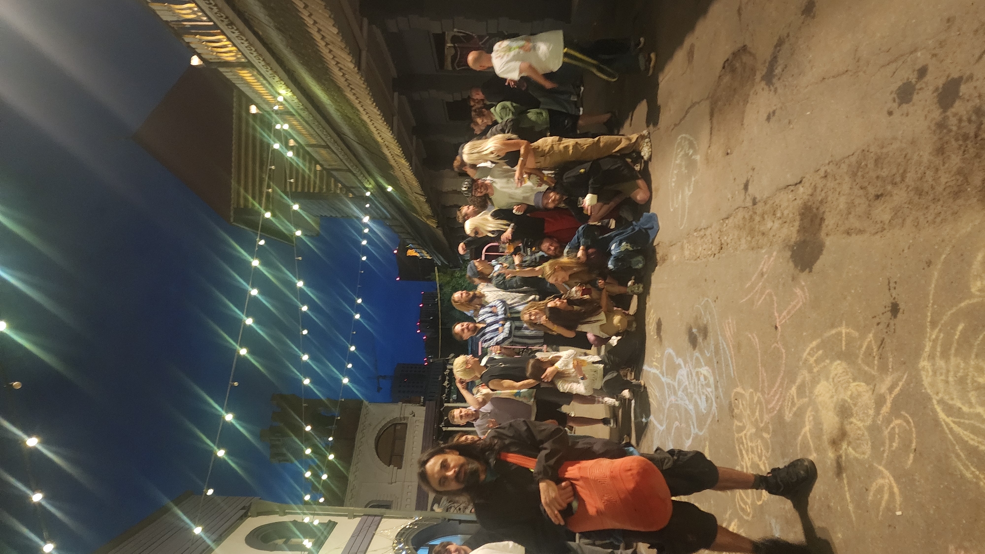 The image depicts a nighttime gathering of approximately 20-30 people in an outdoor urban setting. The scene appears to be a public space, possibly a street or alleyway, adorned with decorative string lights overhead. The architecture suggests a historical or themed area, with buildings featuring decorative elements and awnings. The ground is paved and shows chalk drawings, indicating a possible artistic or community event.
In the foreground, a man with dark hair and a beard is standing, wearing dark clothing and carrying an orange tote bag over his shoulder. He is looking directly at the camera. Behind him, a diverse group of individuals, both men and women of various ages, are congregated. Some are standing, while others are sitting or kneeling on the ground. They are dressed in casual attire. Several people are holding drinks and appear to be engaged in conversation and social interaction. The atmosphere seems relaxed and convivial. The background shows more buildings and the dusky night sky. Based on the architectural style and the presence of such gatherings, this scene is likely located in the **Red October (Krasny Oktyabr) district of Moscow, Russia**, a former industrial zone now transformed into a hub for creative industries, bars, restaurants, and cultural events, often featuring outdoor gatherings and artistic displays.