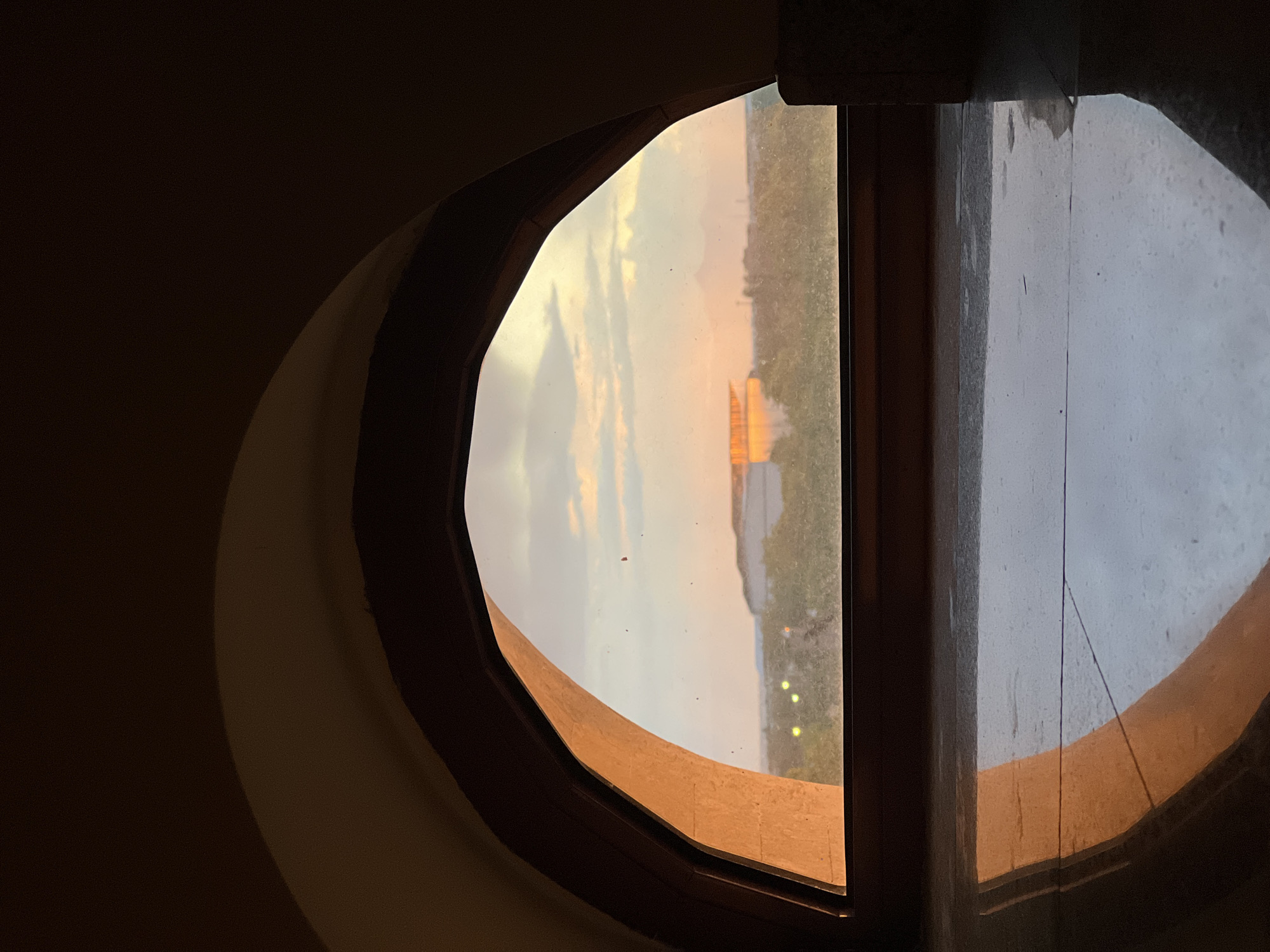 A circular window frames a view of Yerevan, Armenia, at dusk. Outside, the landscape includes greenery, a river, and a distant golden structure, possibly a building or monument, illuminated by the setting sun. Lights are also visible along the riverbank. The windowpane has some smudges and dust. The interior wall is dark. The window frame is made of light brown material.