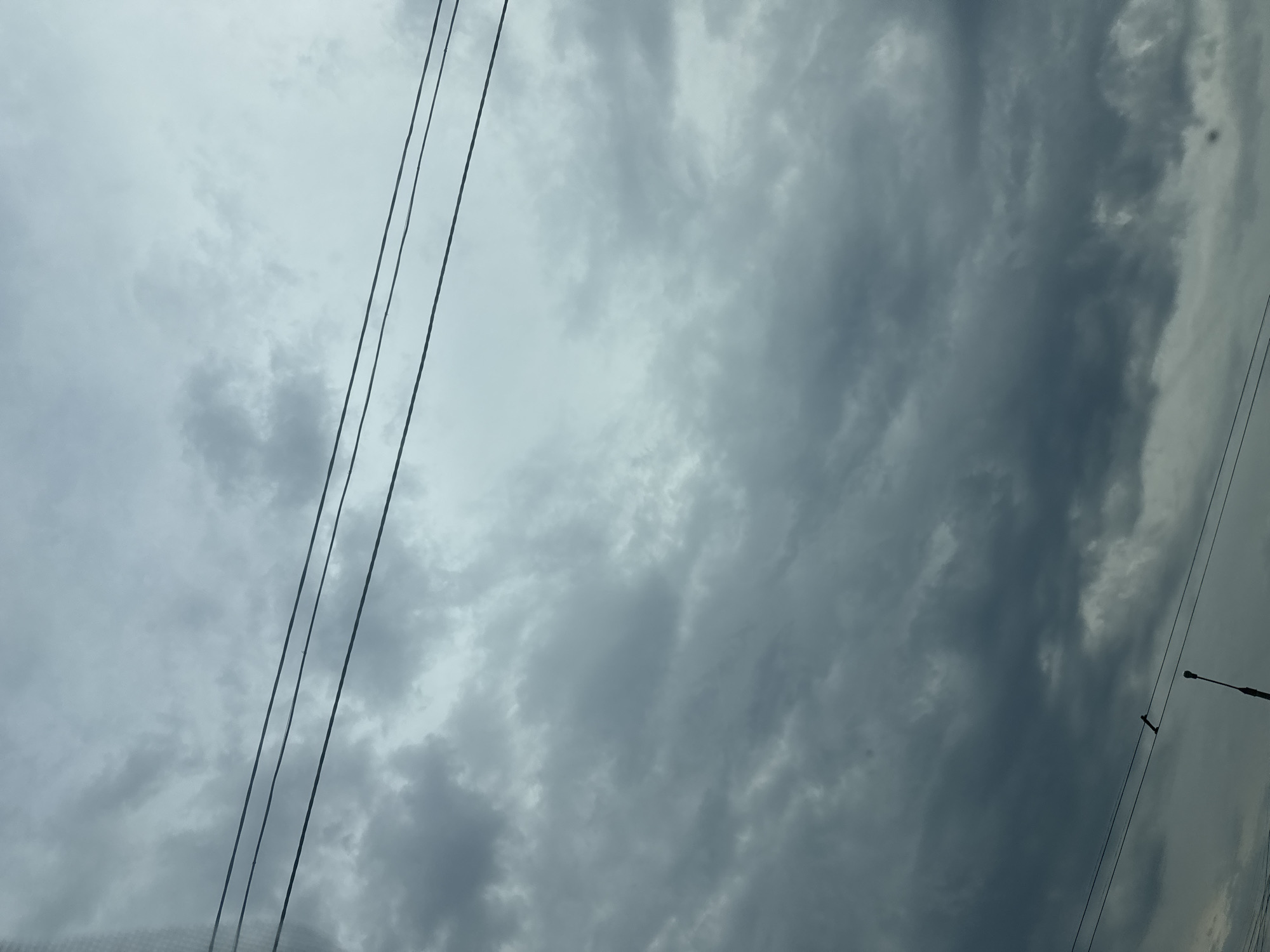 The image presents an extensive view of a heavily overcast sky, characterized by varying shades of grey and desaturated blue clouds; lighter tones are visible in the upper sections, transitioning to denser, darker formations toward the bottom. Three dark, parallel utility lines horizontally traverse the upper-middle portion of the frame. In the lower-middle and right, the silhouette of the upper section of a dark street light pole is discernible. An elongated, dark, angled object appears to be attached to or hanging from the lowest utility line, positioned slightly to the left of the street light. Faint grid patterns are present along the far-left edge of the image, indicating the photo may have been captured through a screened opening. The overall illumination is diffuse, with no direct light source visible. There are no identifiable people, animals, or vehicles within the scene, which is situated in the city of Unknown, Unknown.