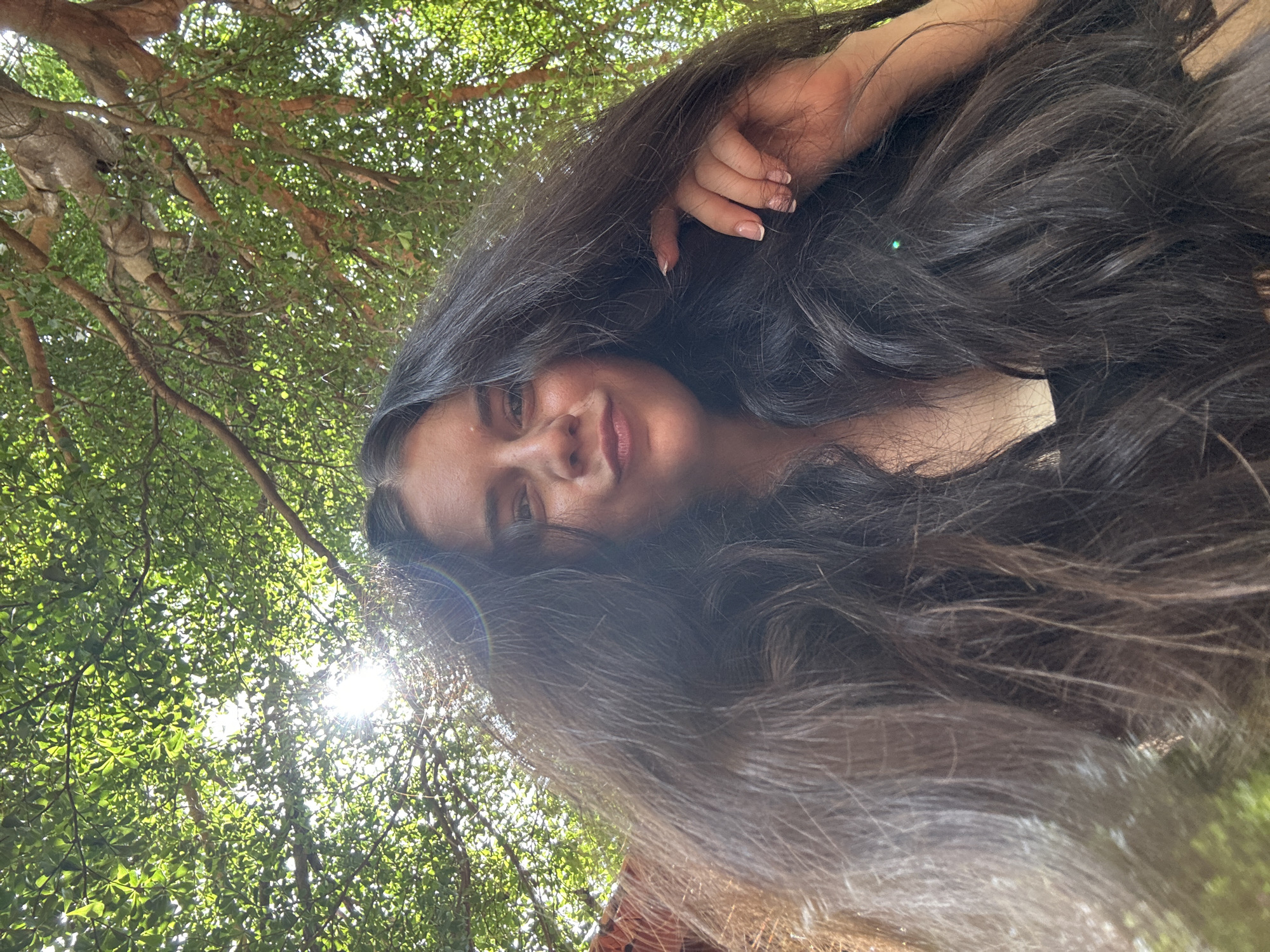 A low-angle selfie shows a young woman with very long, dark, wavy hair that appears to be blowing in the wind, obscuring much of her face. She is looking up towards the camera with a slight smile and her eyes are visible. Her right hand is raised and touching her hair, with a faint green glint visible on her hair. The background is dominated by the dappled green leaves and branches of a tree, with sunlight filtering through. A portion of what appears to be an orange fabric is visible on the left edge of the frame. The scene suggests an outdoor setting, possibly in a park or garden in Mũi Né, Vietnam, given the foliage and the general lighting conditions.