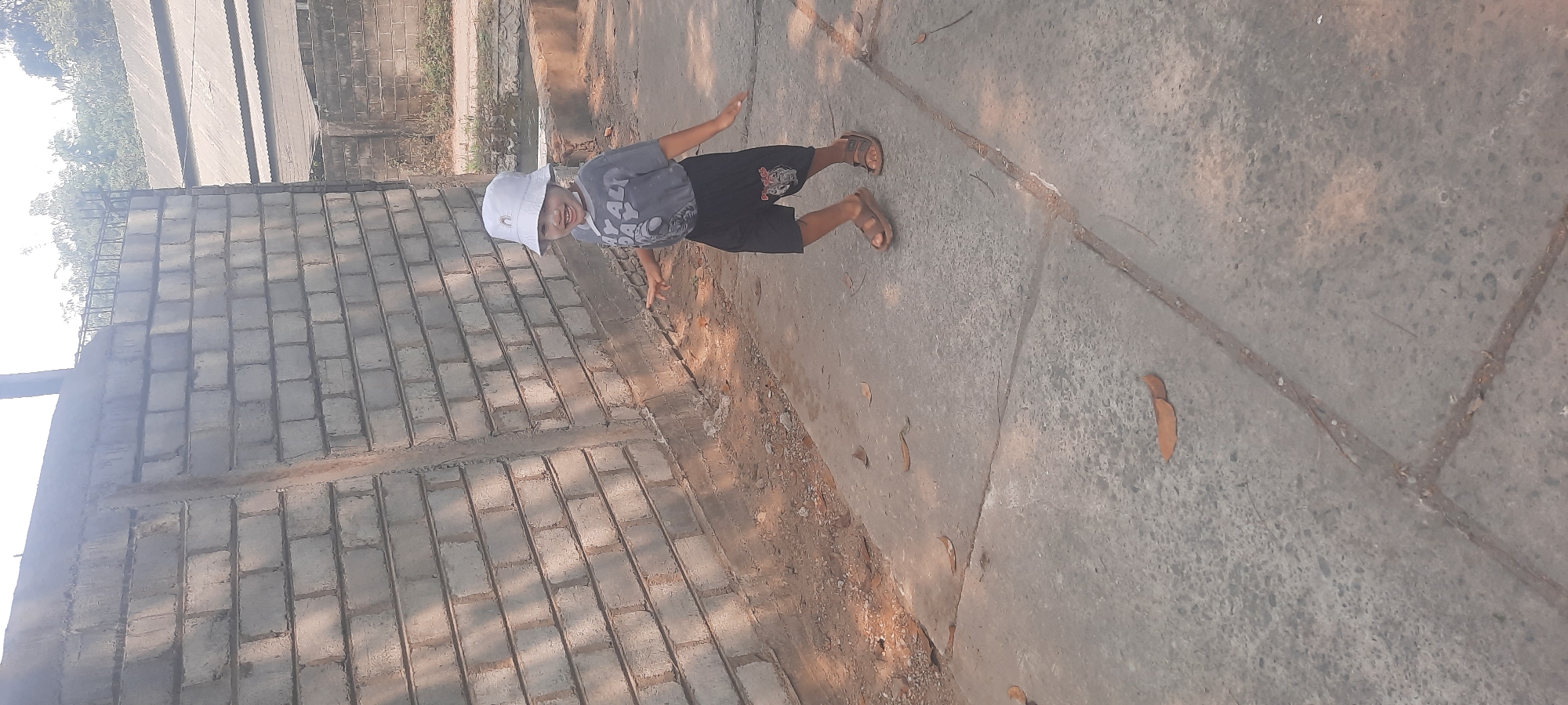 A child wearing a white bucket hat and a grey t-shirt with dark shorts is running on a concrete path. The child is smiling and looking towards the camera. The child's right hand is extended to the side, and the left hand is slightly raised. The child is wearing brown sandals. A grey brick wall is to the left of the child, and dry leaves are scattered on the ground. In the background, there are some trees and buildings with corrugated metal roofs. The scene appears to be an outdoor environment in Ploso, Indonesia, likely a residential area or an undeveloped plot of land.