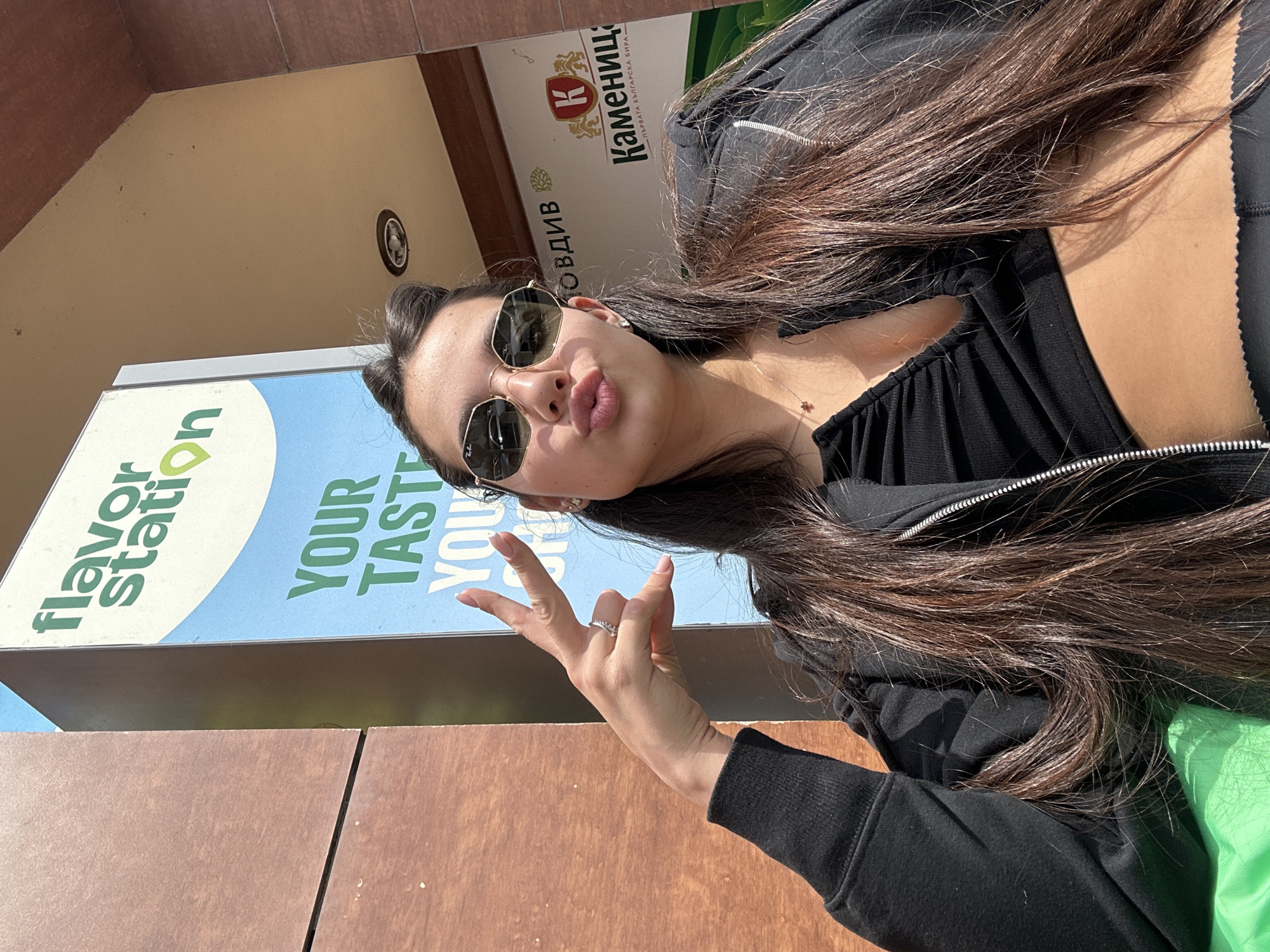 A young woman poses outdoors in Plovdiv, Bulgaria. She wears sunglasses and dark clothing. She is making a hand gesture. Behind her are signs in Bulgarian, including one for "Каменица" beer and another that says "Flavor Station." The background includes beige-colored walls.