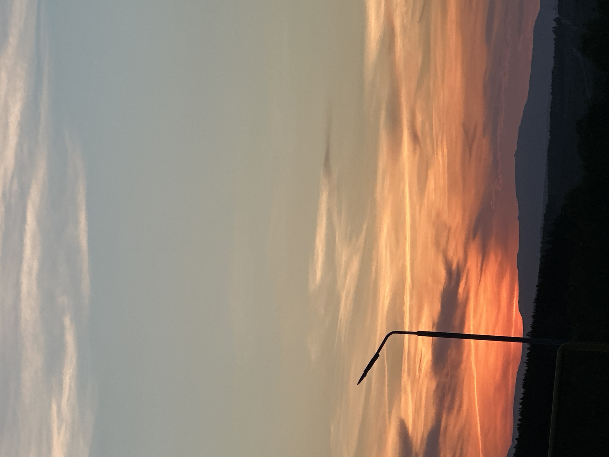 Here's a description of the image:
The image depicts a sunset sky. The right side of the frame is dominated by vibrant hues of orange and yellow, transitioning into softer shades towards the center. Wispy clouds are visible in the orange and yellow sections, mirroring the cloud formation on the left, where the sky is a pale blue. A dark silhouette of a hill or mountain range is seen on the far right, and a street lamp is prominently featured in the foreground. The photo's location is in the city of Unknown, Unknown.