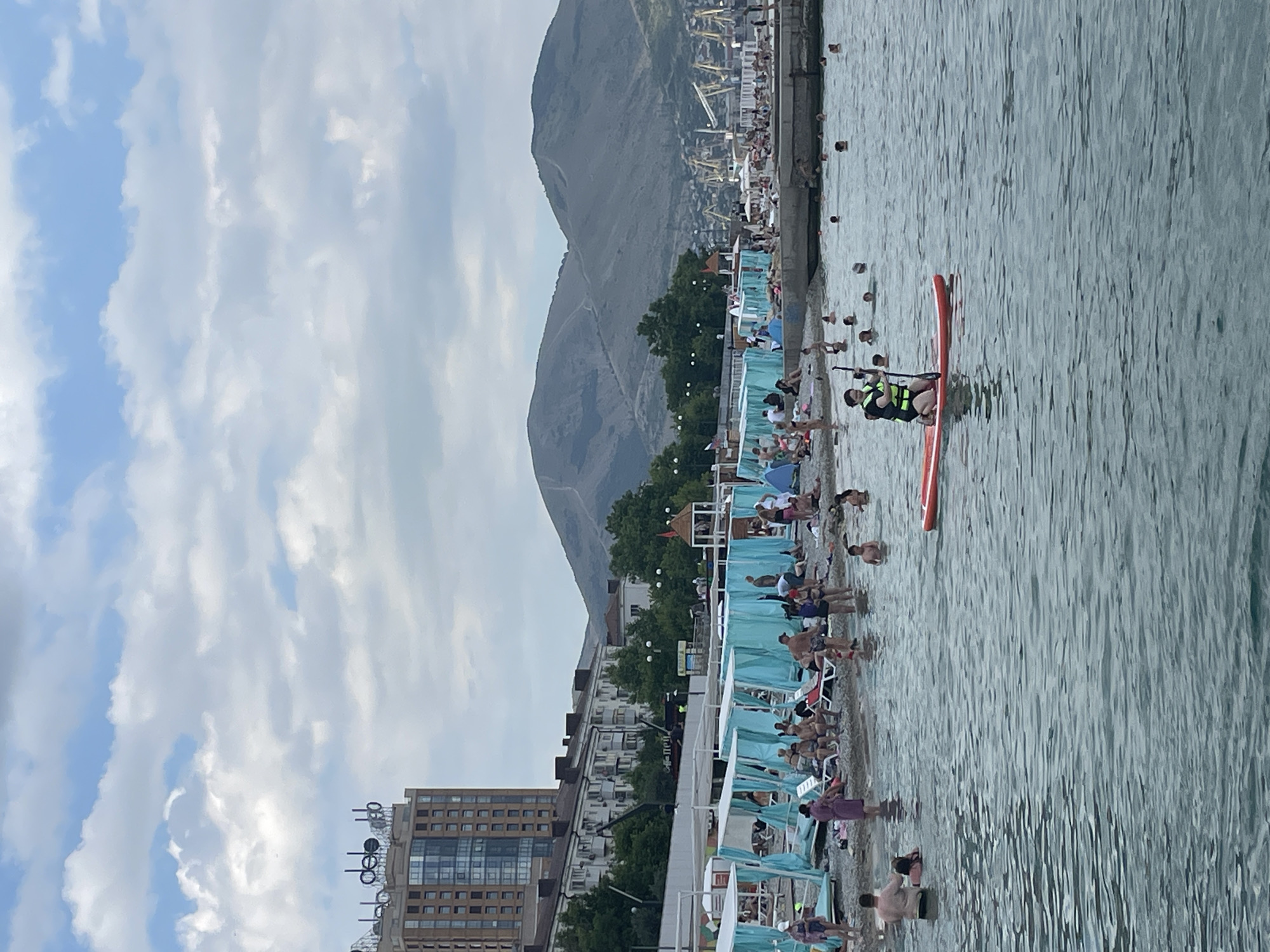 The image depicts a busy beach scene in Novorossiysk, Russia. A multi-story building with "CEEL" signage on its roof is visible in the background, along with apartment buildings and a mountainous landscape. The beach is lined with rows of turquoise beach tents and umbrellas, with many people relaxing on lounge chairs. In the water, several individuals are swimming, and one person is paddleboarding. The sea is calm, and the sky is partly cloudy. The overall atmosphere is one of a typical summer day at a crowded seaside resort.