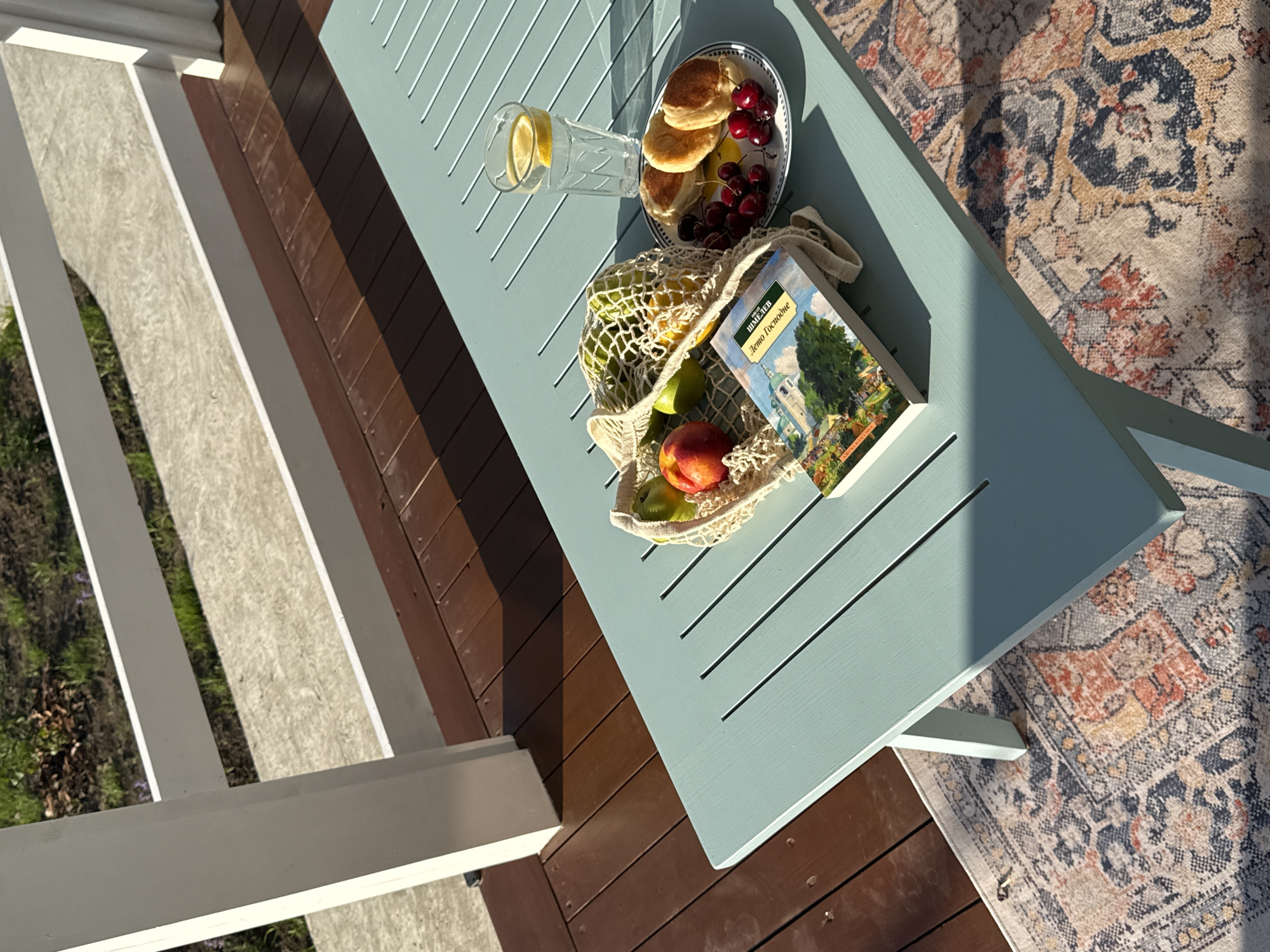 A high-angle shot captures a light blue slatted table on a wooden deck, bathed in dappled sunlight. The table holds breakfast items: a plate of golden pancakes topped with cherries and a glass of water with lemon slices. Beside these, a mesh bag overflows with fresh fruit, including apples, limes, and a nectarine. A book with a colorful cover featuring a village scene and a large tree rests on the table, its title clearly visible.
The background shows a patterned rug with warm, earthy tones and a hint of white railing bordering a grassy area. The scene suggests a leisurely morning or afternoon outdoors, perhaps on a porch or patio. The strong shadows indicate it's either early morning or late afternoon. The presence of breakfast food and a book implies a moment of relaxation and quiet enjoyment. The text on the book cover is in Cyrillic and reads "Лето Господне" (Leto Gospodne), which translates to "The Lord's Summer."