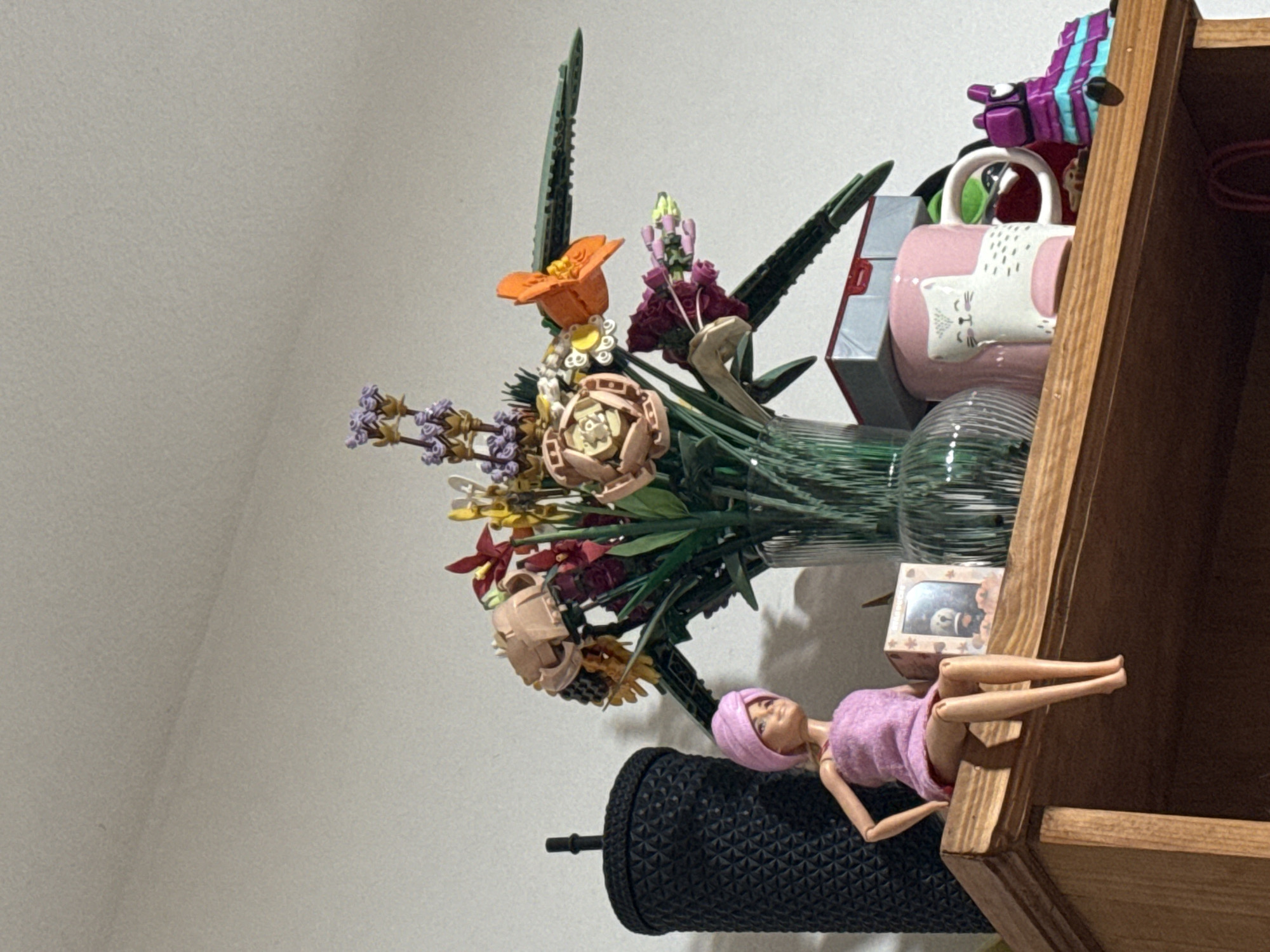 On a wooden shelf against a white wall, sits a black textured tumbler supporting a Barbie doll wearing a pink outfit and head covering. Adjacent to the Barbie doll is a small picture frame and a clear, ribbed vase containing an arrangement of LEGO botanical flowers. A pink mug with a white cat design and various other items, including LEGO blocks, are also positioned on the shelf. The setting is an interior space. There is no information available in the image to determine its location in Puebla, Mexico.