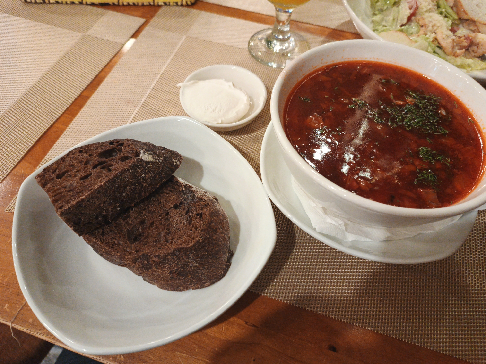 This image depicts a meal at a restaurant in Bac Nha Trang Ward, Vietnam. The table is set with a woven placemat and a wooden table. On the placemat, there is a white bowl filled with a deep red soup, garnished with chopped green herbs. To the left of the soup bowl, a smaller white bowl contains a dollop of white cream, likely sour cream or yogurt. In front of the soup and cream, a white, curved dish holds two slices of dark, dense bread. In the background, a white bowl contains a salad with lettuce, tomatoes, and croutons. A glass with an orange liquid and a silver-colored object, possibly a wine glass or a utensil, are partially visible at the top of the image. The lighting suggests an indoor setting, possibly a restaurant, with natural light from a window or overhead lights illuminating the food.