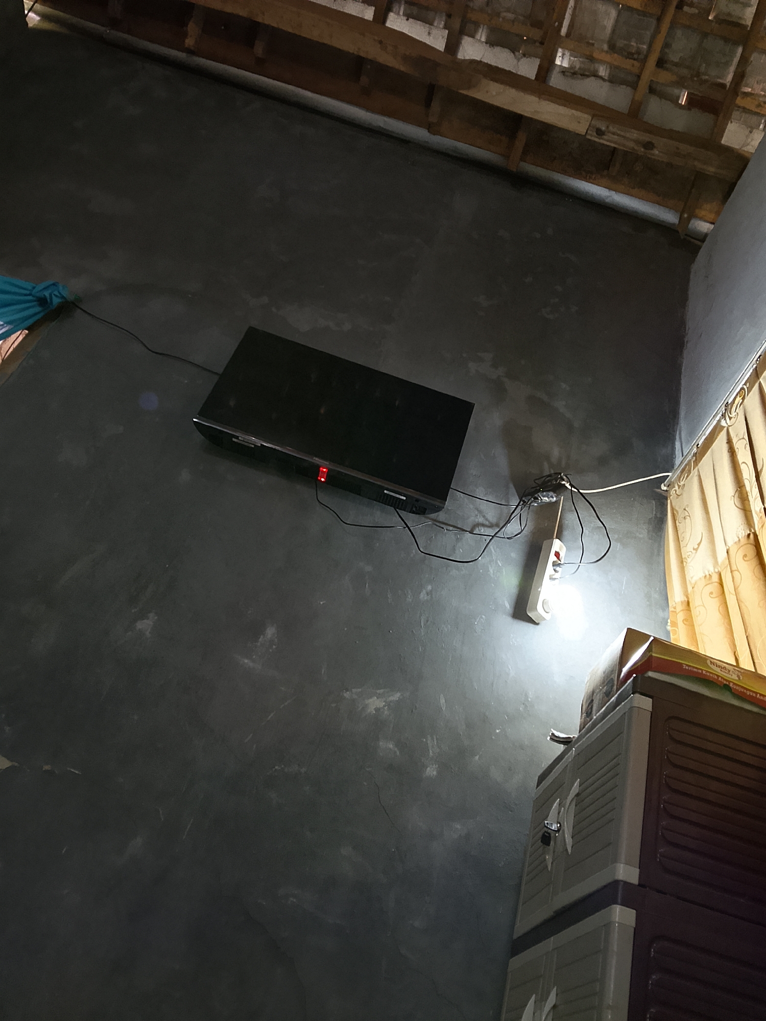 The image depicts an interior scene, likely a modest dwelling, in Besuki, Indonesia. A black television set is mounted on a dark gray wall. To the right of the television, a white electrical outlet is visible, with a light bulb screwed into it, illuminating the area. Several black electrical cords are connected to the outlet and snake across the wall. In the lower right quadrant of the image, a two-tiered brown and gray plastic cabinet is partially visible, with a red and yellow cardboard box resting on top. Yellow curtains hang on the far right side of the image. The ceiling appears to be constructed with wooden beams and what looks like terracotta tiles. The overall lighting is dim, with the primary light source emanating from the bulb in the outlet. There are no people or animals visible in the frame.