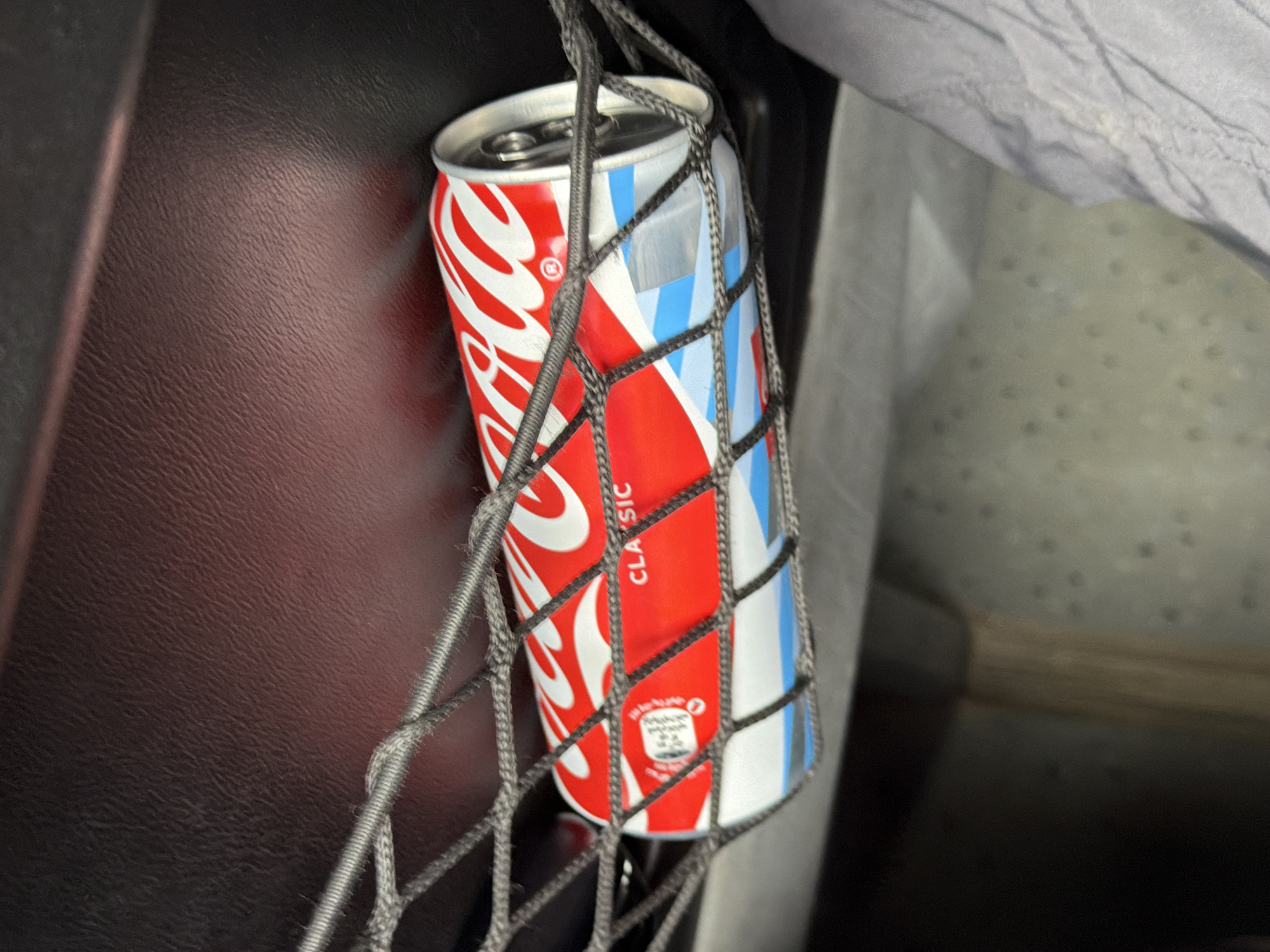 A Coca-Cola Classic can is secured in a black mesh net pocket, likely located on the back of a car seat. The can is red and white with the Coca-Cola logo and white wavy stripes. Part of the can's label, showing nutritional information, is visible on the left side. The background consists of a dark, textured surface, possibly leather or plastic, and the edge of a light-colored fabric, potentially clothing or another seat. The lighting is subdued.