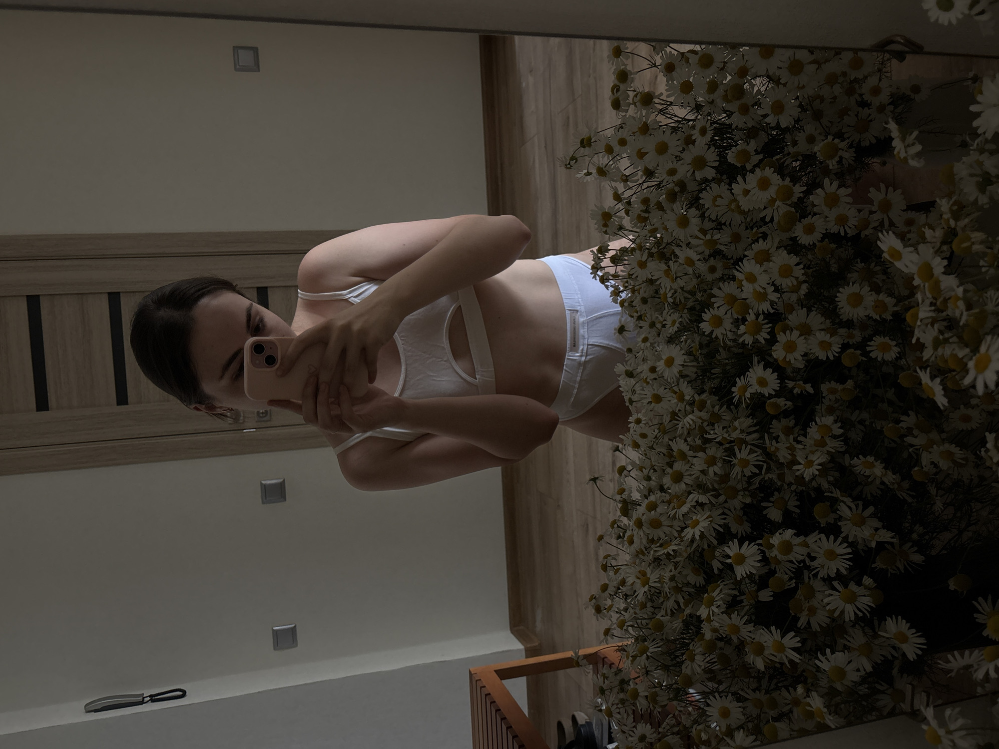 A woman in white lingerie is taking a mirror selfie. She is wearing a white bralette and matching white high-waisted underwear. Her arm is extended as she holds up a pink phone to capture the reflection. The background of the image shows a room with light-colored walls and wooden furniture. A large bouquet of white daisies is prominently displayed to the right of the woman, adding a touch of nature to the scene. The overall lighting suggests it might be daytime, perhaps late afternoon, given the soft shadows. The woman's expression is neutral as she focuses on taking the photo. The reflection in the mirror also shows a wooden door and wall decor, suggesting a domestic setting.
