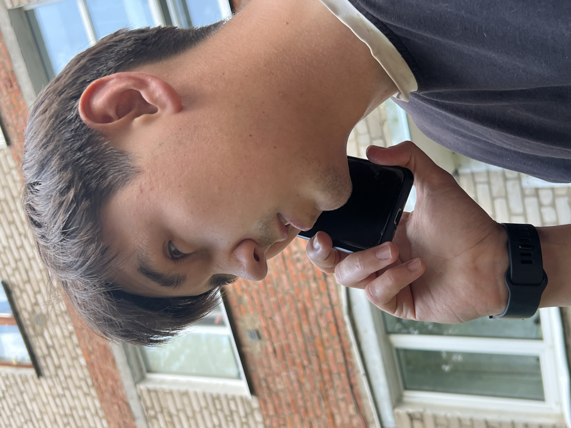 A young person is captured in a close-up, side-profile shot, holding a black smartphone to their ear. They are wearing a dark t-shirt with a white collar. A black smartwatch is visible on their left wrist. The background features a brick building with windows and hints of greenery. The lighting suggests it is daytime, possibly overcast, as there are no strong shadows. The person's expression is neutral, focused on the phone conversation. The scene appears to be outdoors, likely in a residential or urban area, given the brick architecture. The orientation of the photo is vertical.