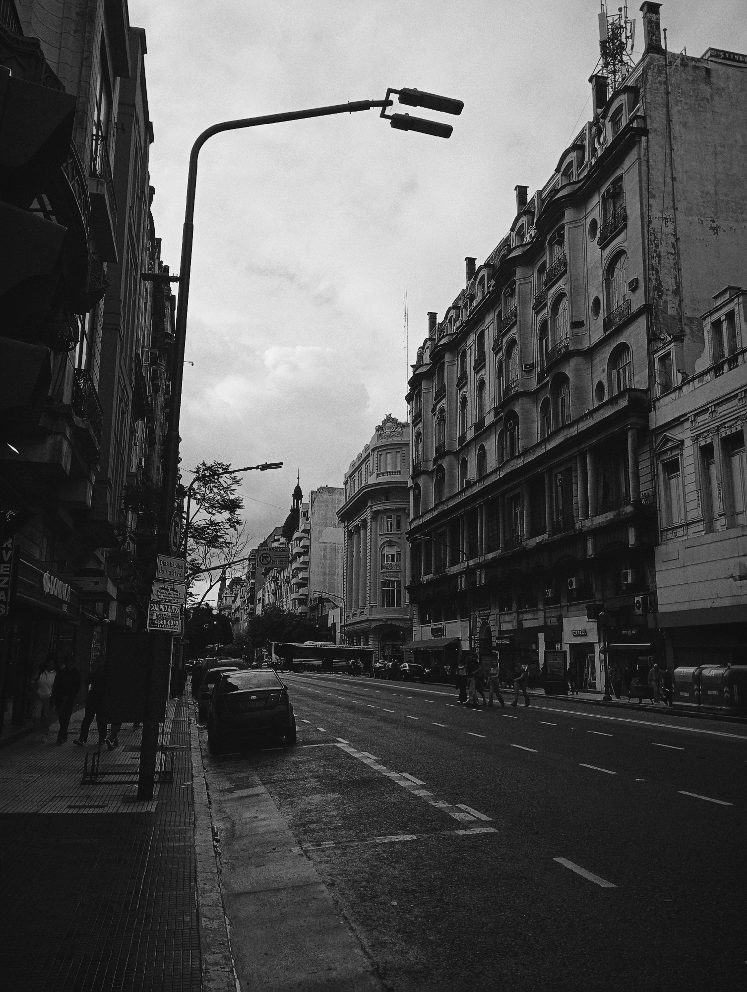 This black and white photograph captures a bustling street scene in Buenos Aires, Argentina, under an overcast sky. The wide asphalt road, marked with white lines, recedes into the distance, flanked by sidewalks on both sides. Tall, ornate buildings with classical architectural details, such as arched windows, balconies, and decorative cornices, line both sides of the street. The building on the right is particularly grand, featuring dormer windows on a mansard roof and a modern antenna array on its highest point.
On the left, a large street lamp with two fixtures extends over the road. Below it, on the sidewalk, several pedestrians are seen walking, mostly in silhouette due to deep shadows. Multiple cars are parked along the left curb, including a dark sedan in the foreground. A vertical street sign attached to a pole on the left sidewalk is visible, with text reading "Días hábiles de 7 a 21 hs." (Working days from 7 AM to 9 PM) and further down, "COMPRO AUTOS 4568-0070" (I buy cars).
Further down the street, a bus is visible, and other cars indicate active traffic. A few people are also seen crossing the street in the mid-ground. Modern trash bins are visible on the right sidewalk. The overall lighting is dim due to the cloudy weather, lending a slightly muted and atmospheric quality to the urban landscape. The scene depicts a typical, busy daytime in a historic city district.