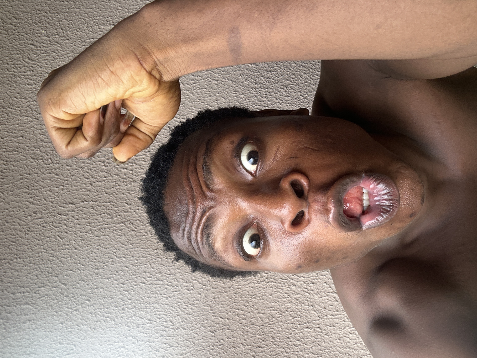The image depicts a person with dark skin, short dark hair, and wide eyes, positioned upside down. The person has an open mouth, revealing their teeth. Their right arm is raised and clenched into a fist near their head. The background appears to be a textured, light-colored wall. The location is unknown.
