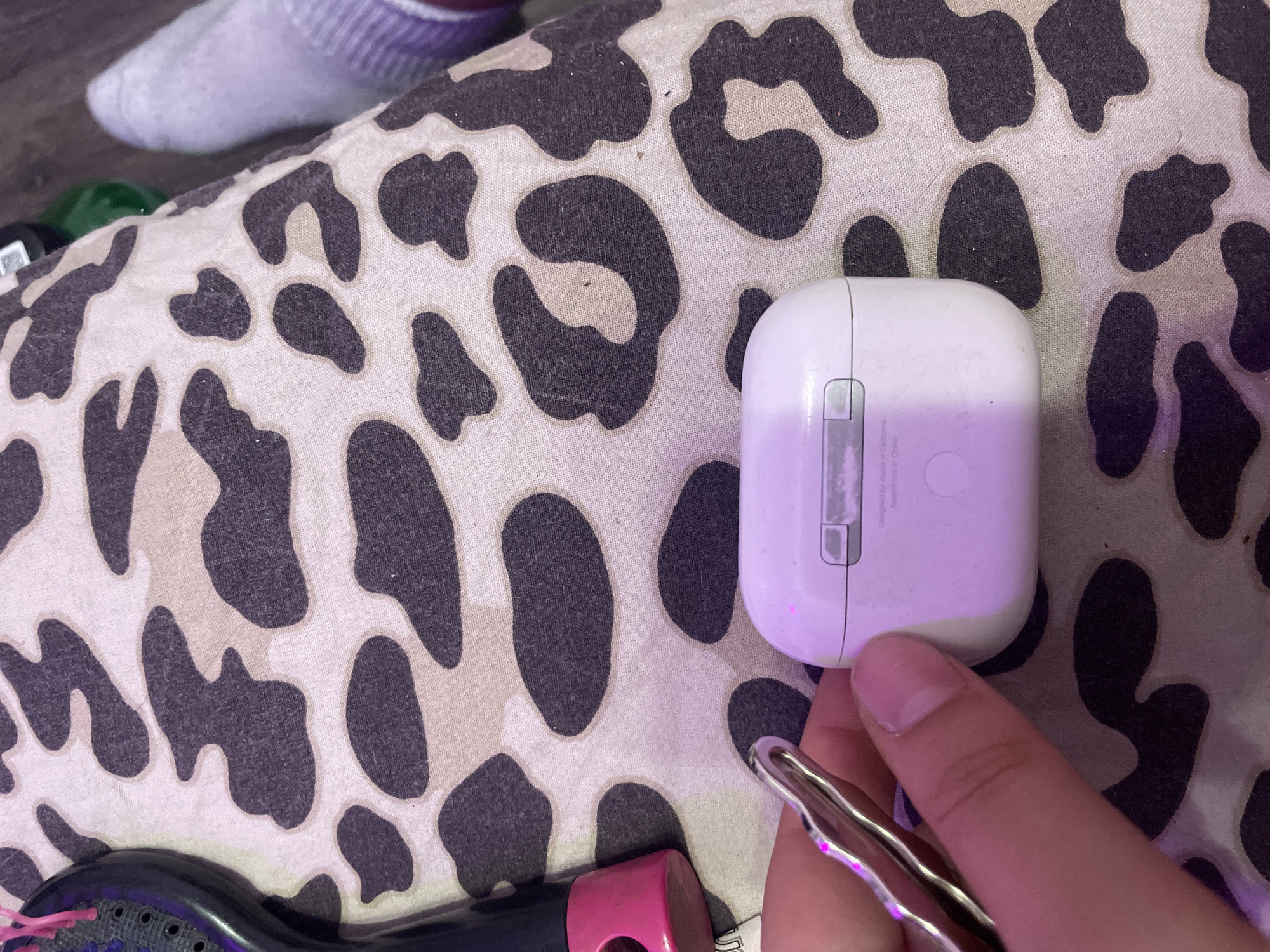 A person's hand is holding a silver keychain near a white AirPods charging case. The charging case is placed on a fabric surface with a leopard print pattern. In the background, a purple brush and a white sock are visible. The scene is lit with purple and pink lighting. This image was taken in Kokkorevo, Russia.