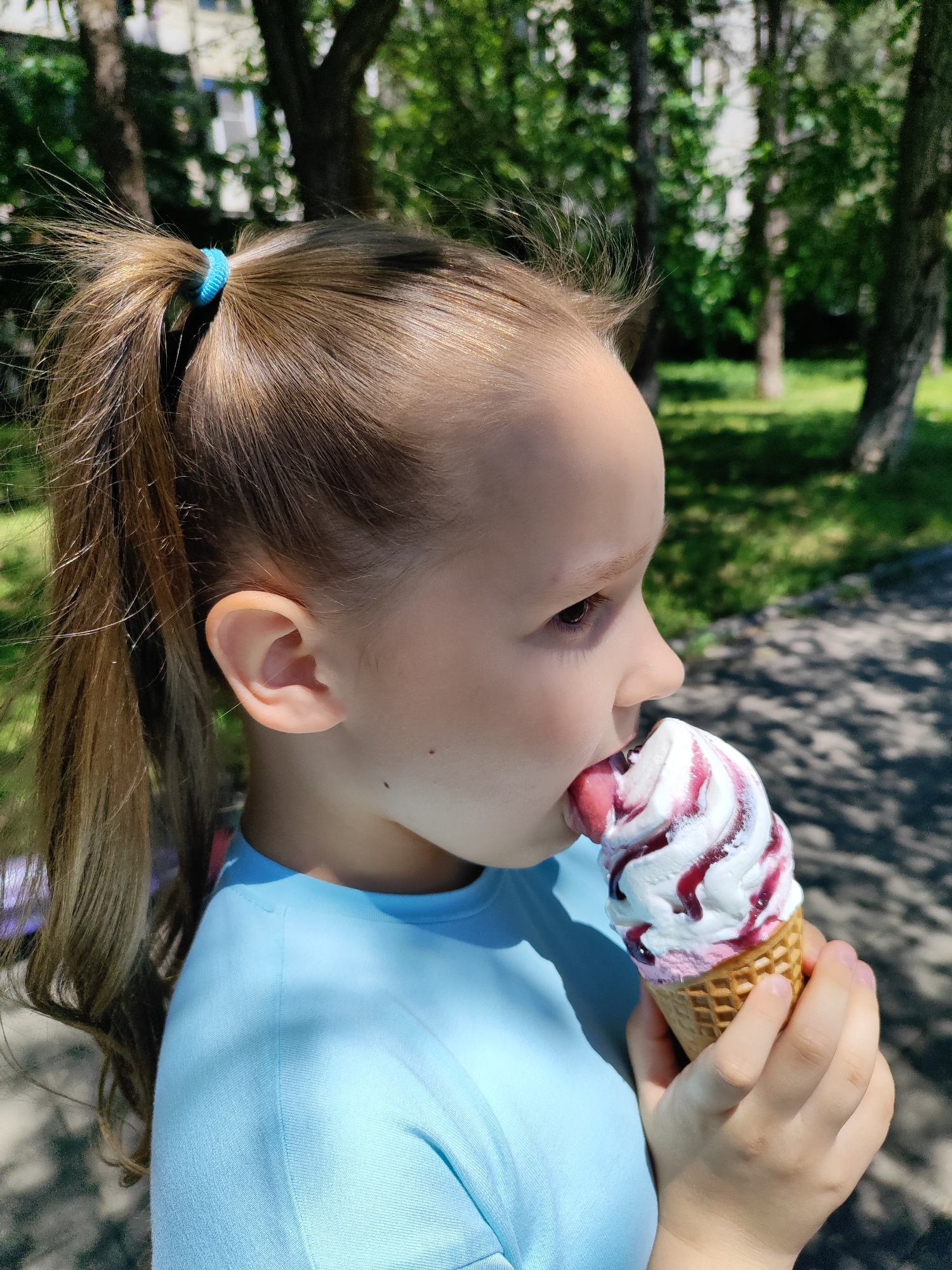 A vibrant, sunlit close-up photograph captures a young child, likely a girl, in profile, enjoying an ice cream cone outdoors in Krasnodar, Russia. The child is positioned on the right side of the frame, facing right, with light brown hair tied back in a high ponytail secured with a bright light blue scrunchie. She is wearing a light blue t-shirt.
Her attention is entirely on the ice cream cone she holds with both hands. The soft-serve ice cream features a white base generously swirled with a reddish-purple syrup, possibly berry or cherry flavored. Her tongue is extended, actively licking the top of the melting treat.
The background is a soft, out-of-focus blur of green foliage and tree trunks, suggesting a park or a tree-lined outdoor setting. Dappled sunlight and shadows fall across the ground and the child's shirt, indicating a bright, sunny day, likely mid-morning or afternoon. A small mole is visible on the child's neck below her ear. No text is visible in the image.