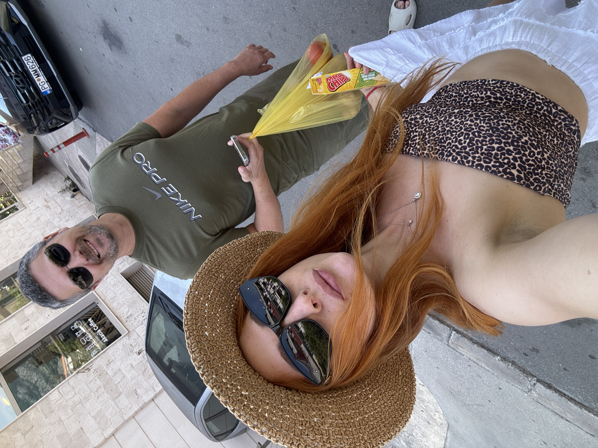 A medium-angle, rotated eye-level shot captures a selfie of two people standing on a street in Budva, Montenegro. A woman with long red hair and a straw hat is in the foreground, wearing sunglasses. She wears a strapless, leopard-print top and a white skirt. She is holding a yellow bag containing a tomato and a yellow package of MEGA CHIPS Sour Cream and Onion chips. A man with gray hair and sunglasses is to the left, wearing a green Nike Pro shirt and holding a cell phone. In the background, there is a car, part of a building with the text "Akacia Coffee" visible on a window, and another black car with the license plate "PG MM 626". The street is made of asphalt.