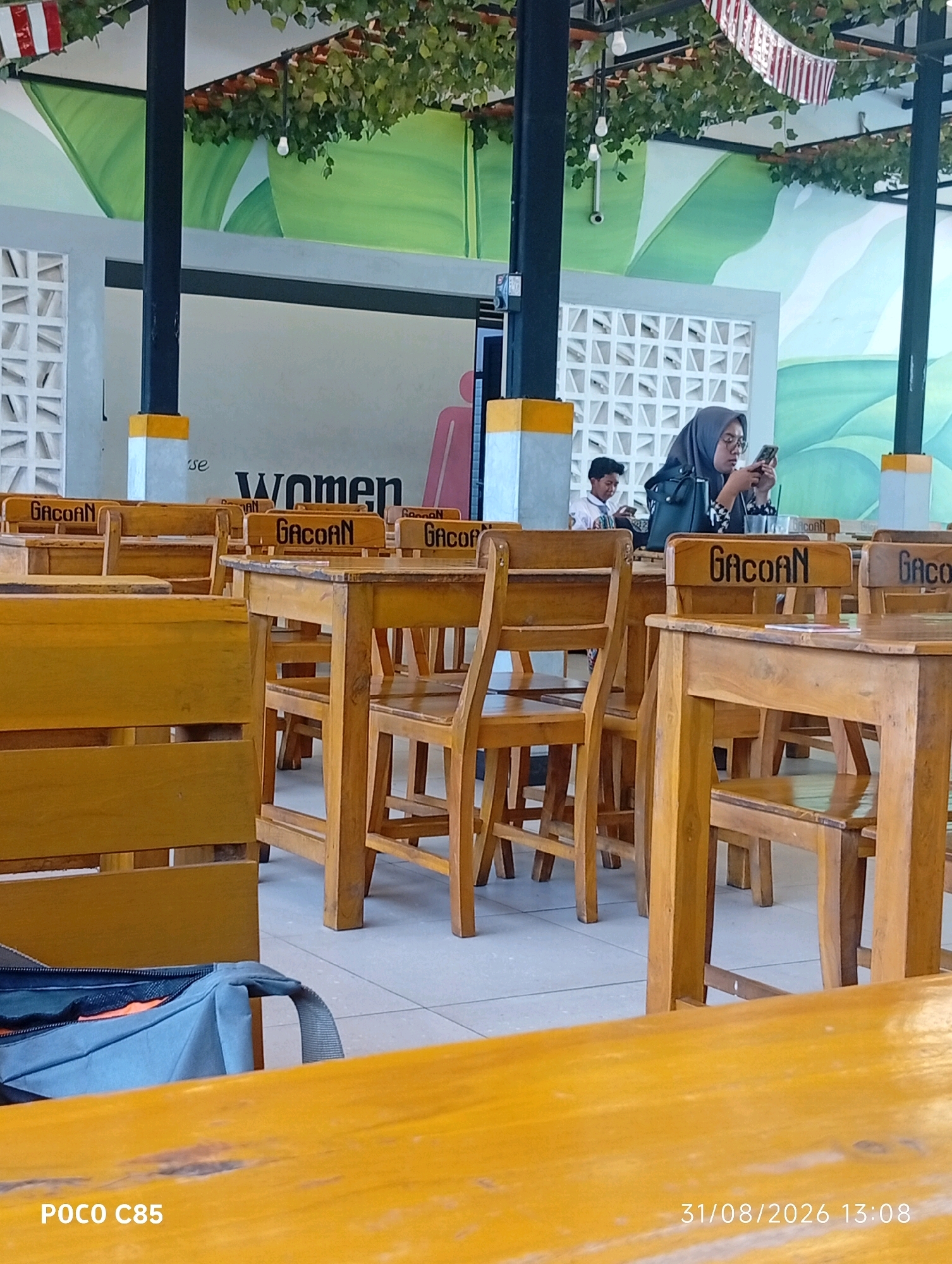 In this medium shot, taken from a slightly low angle, a busy seating area within an outdoor or semi-outdoor cafe is depicted. The foreground is dominated by a bright yellow-orange wooden table, with a blue bag resting on a similar colored surface to its left. Beyond this table, several rows of wooden tables and chairs are visible, creating a sense of depth. Each chair back is branded with the word "GACOAN" in black lettering.
In the mid-ground, a woman wearing a gray hijab and glasses is seated at one of the tables, engrossed in her smartphone. She is dressed in a dark jacket with a patterned blouse. Beside her, a young boy in a white shirt is also looking at a device. The background reveals a mural painted on a wall, featuring large green leaf motifs and a white decorative wall section. Red and white striped pennants are strung across the ceiling structure, adorned with artificial greenery and a hanging light bulb.
The environment appears to be a public establishment, possibly a cafe or restaurant, given the arrangement of tables and chairs. The presence of the "GACOAN" branding suggests it might be a franchise or a location associated with that name. The overall impression is one of casual dining or meeting space. The timestamp "31/08/2026 13:08" and "POCO C85" are visible in the bottom right and left corners respectively, indicating the date, time, and the device used to capture the image. Based on the branding and the visual cues, the location is likely a "Kedai GACOAN" restaurant in South Tangerang, Indonesia, known for its unique branding and often found in such casual, open-air settings.