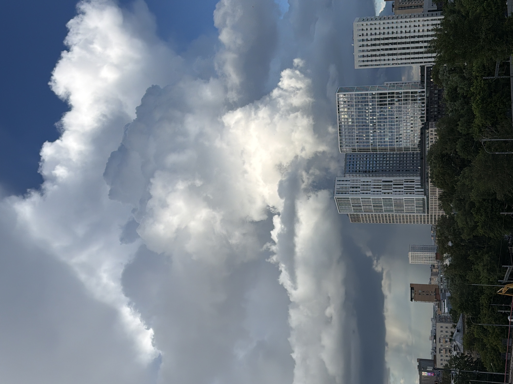 This media file shows a dramatic sky filled with large, billowing white and gray clouds, contrasting with a patch of clear blue sky. In the foreground, several modern, multi-story buildings with distinctive grid-like facades are visible, some appearing to be residential or office complexes. Trees with lush green foliage line the right side of the frame, obscuring parts of the buildings and the background. The lighting suggests it could be daytime, possibly late afternoon, with the sun illuminating some of the clouds and reflecting off the windows of the buildings. The overall scene conveys a sense of urban development set against a dynamic natural sky. The orientation of the image is vertical, implying it was captured in portrait mode. No people are clearly visible, and there is no discernible text in the image. The weather appears to be changing, with the heavy clouds possibly indicating an approaching storm or a recent downpour.