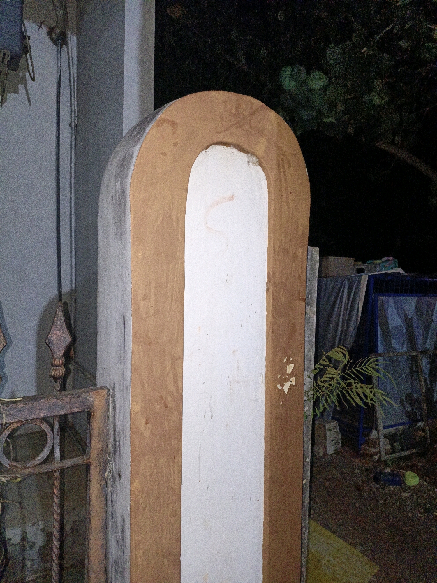 The image shows an outdoor scene at night, possibly in the city of Cikal, Indonesia. In the foreground, a tall, arched structure made of concrete and painted white and brown stands prominently. To the left of the structure, a rusty metal gate with circular and decorative elements is partially visible, along with a black metal pole extending upwards. The background is dark, with foliage from trees and shrubs visible to the right, and some structures covered with tarpaulins. The ground appears to be covered with gravel and dirt, with some scattered debris. There are no people or animals in the image. The overall impression is of an outdoor area with some construction or decorative elements, likely in a residential or public space.