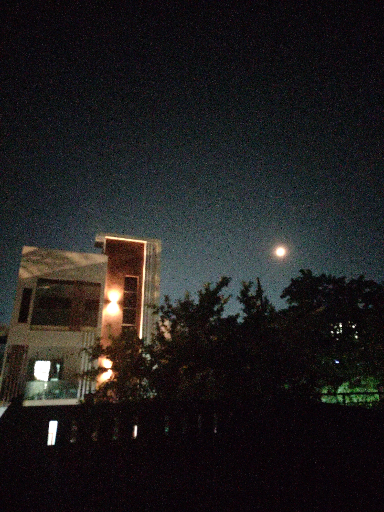 The image depicts a modern building at night, possibly in the city of Bhadrachalam, India. The building has several floors with large windows, some of which are illuminated from within. A prominent feature of the building is a vertical strip of bright light running up its facade. In the background, silhouetted trees and the night sky are visible. A bright, full moon is positioned in the upper right quadrant of the sky. The foreground is dark, with a fence-like structure and some illuminated rectangular shapes suggesting windows or lights. The overall atmosphere is serene and nocturnal.