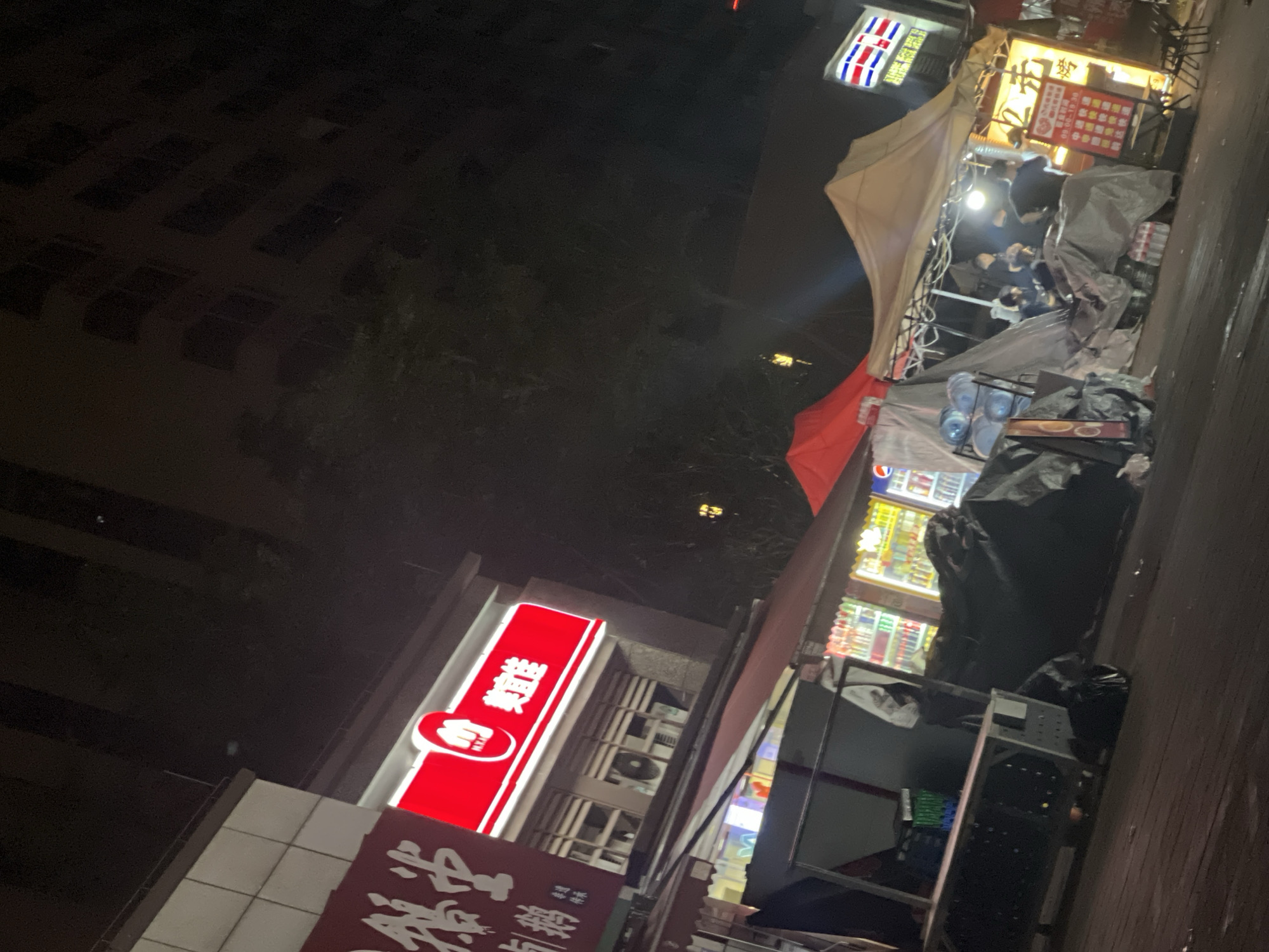 The image captures a vibrant night street scene, likely a bustling market or food street in Chongqing High-tech Zone, China. The scene is dominated by brightly lit temporary stalls and storefronts against a dark, nocturnal sky, suggesting late evening or night.
In the middle ground on the right, a large, light-colored tent structure shelters several individuals. People are gathered under this tent, some partially visible, appearing to be either vendors or customers interacting at a food or retail stall. Bright lights illuminate the area beneath the tent, highlighting activity. Surrounding the stalls are various objects, including stacks of plastic water bottles, coolers displaying drinks, and what appear to be bundled items or trash bags. A large red umbrella is partially visible near the tent.
To the left, a multi-story building rises into the dark sky, with some visible windows, suggesting an urban residential or commercial structure. Below this building, a prominent, vertical red sign with white Chinese characters glows brightly. The most legible character is "汇" (huì), which can mean "to gather" or "to assemble." Below this character is a white emblem resembling a fist or a hand holding something. Another red sign, partially obscured, is visible lower down, also displaying Chinese text. Further right, another brightly lit sign features a red and white patterned design, possibly a flag or logo, with some Chinese characters including "味" (wèi - taste) below it. Above one of the market stalls, a yellow-lit signboard displays multiple Chinese characters and numbers, likely a menu with prices.
The overall impression is one of a lively urban night market or commercial area, with people congregating and businesses operating after dark.