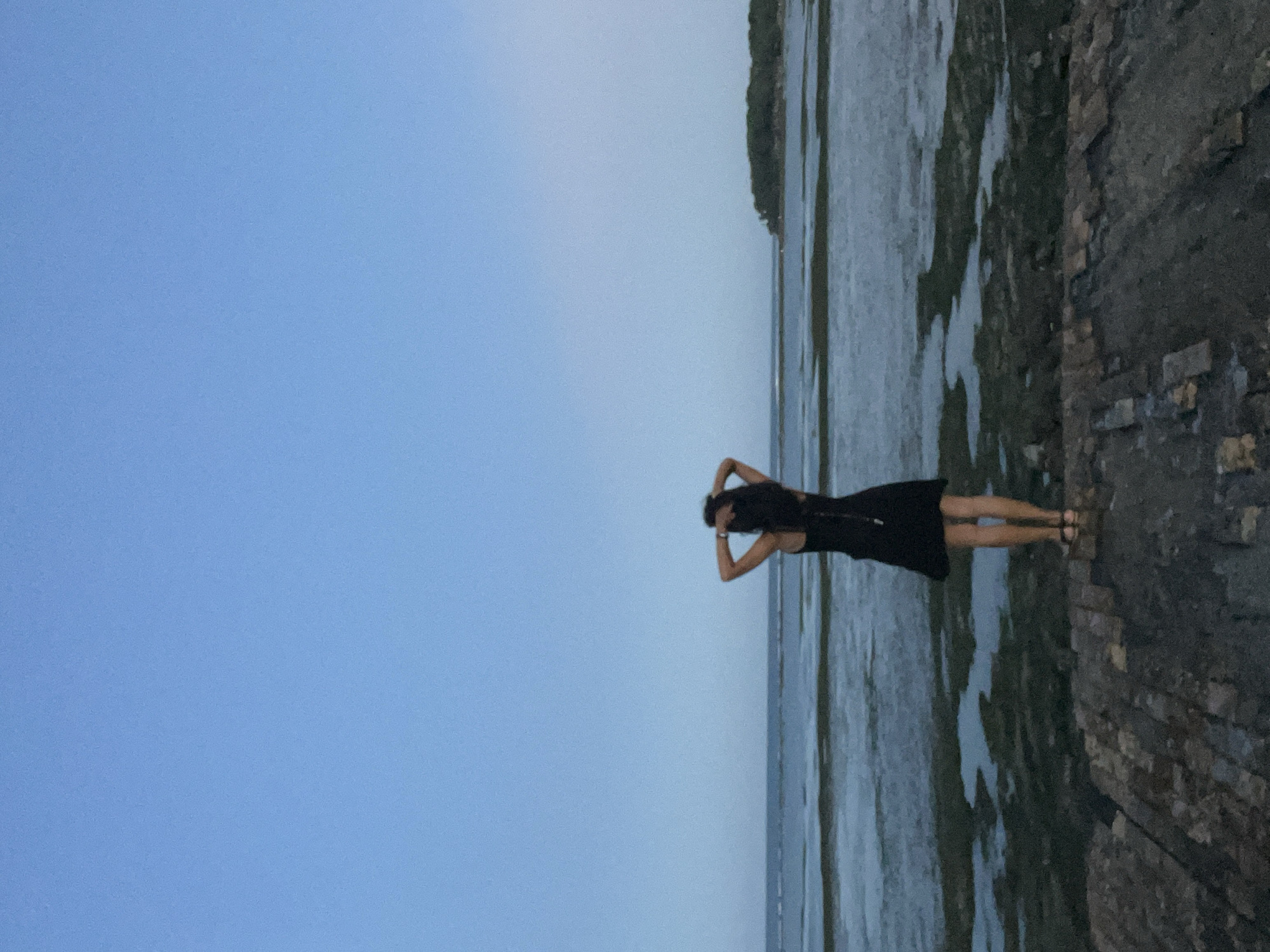 A photograph shows a woman standing on a rocky wall at low tide in Kuta Selatan, Indonesia. The woman is wearing a black dress and sandals, and her arms are raised with her hands behind her head. To the right of the wall, the water recedes to reveal a surface covered with seaweed. Behind the water, there is vegetation on the land. To the left of the woman is a large area of light blue sky.