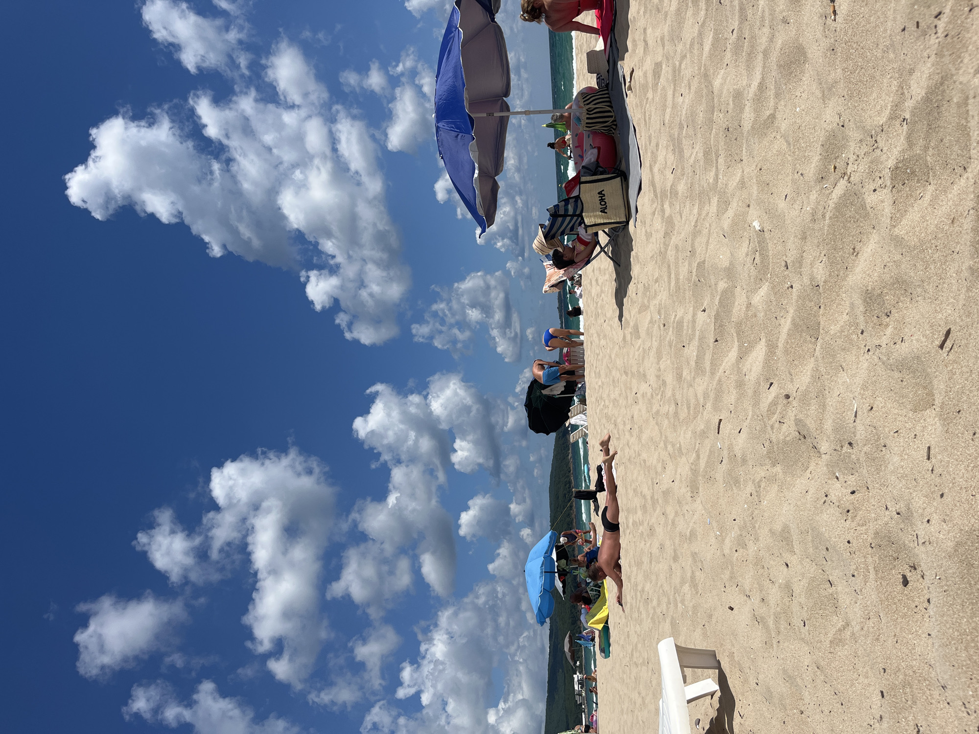 This is a photograph of a beach in Primorsko, Bulgaria, on a sunny day with a blue sky filled with scattered white clouds. The foreground shows a sandy beach with ripples and some debris. Several people are present, enjoying the beach. In the center foreground, a man is lying face down on the sand wearing black swim trunks. To the left, a white lounge chair is partially visible. Further back, a blue umbrella shades a group of people sitting on the sand. To the right, a larger blue umbrella shades more beachgoers, including a woman sitting in a chair with an "ALOHA" bag. Other umbrellas, lounge chairs, and people are scattered across the beach and in the shallow water in the background. The sea stretches to the horizon, and a green, tree-covered hill forms the backdrop.