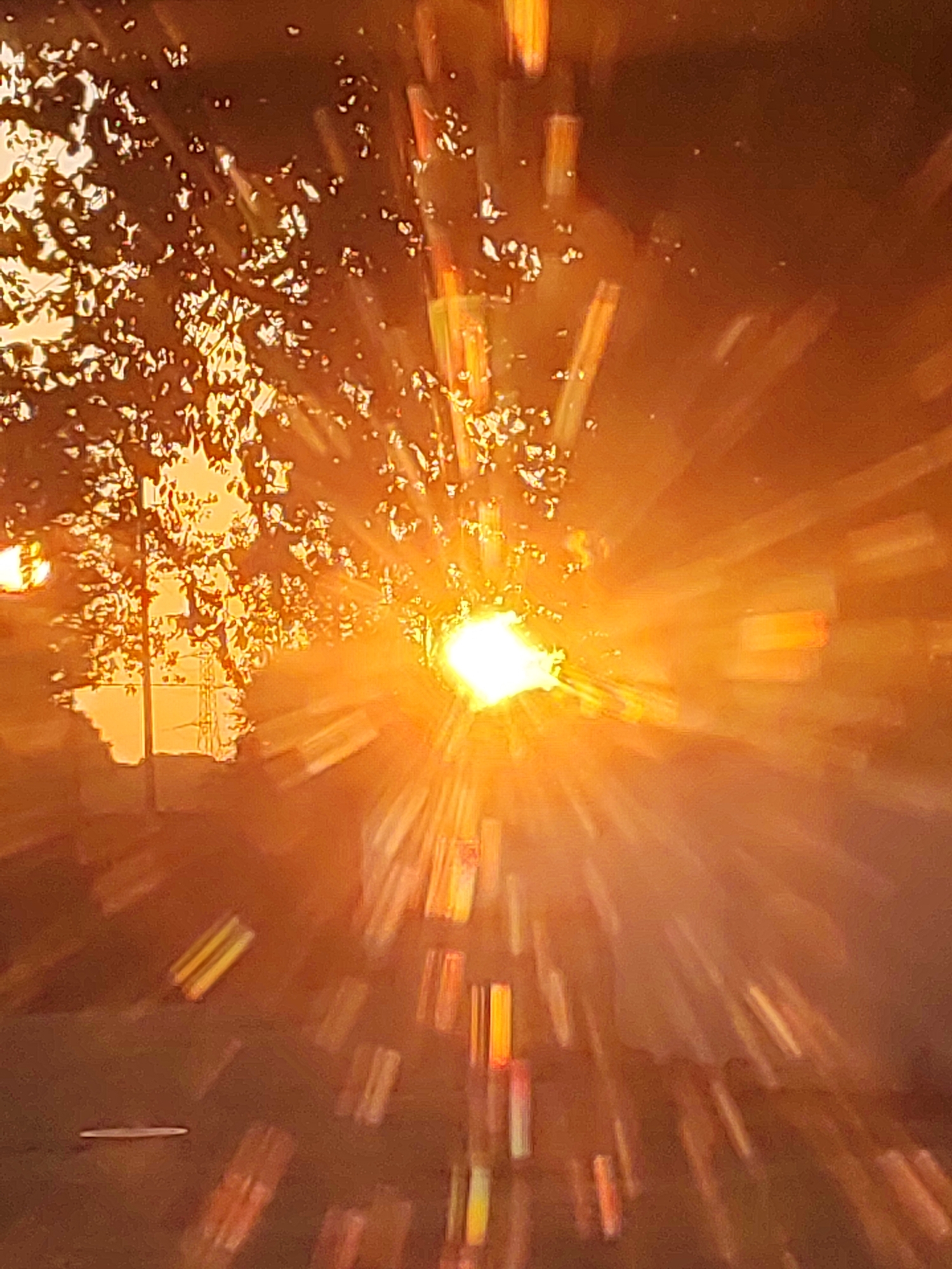 The image shows a sunburst effect, likely caused by the sun shining through foliage or a window with blinds. The sun is bright white and is radiating light outwards in all directions, creating streaks of light. The background is blurred and appears to be dark brown with areas of orange and yellow light. In the upper left portion of the image, there are dappled shadows that suggest the presence of leaves. In the lower part of the image, there are rectangular shapes that appear to be light reflections or glints. The overall impression is of a bright, atmospheric scene. The location is Traspinedo, Spain, but there are no discernible landmarks to confirm this specifically within the image itself, beyond the provided metadata.