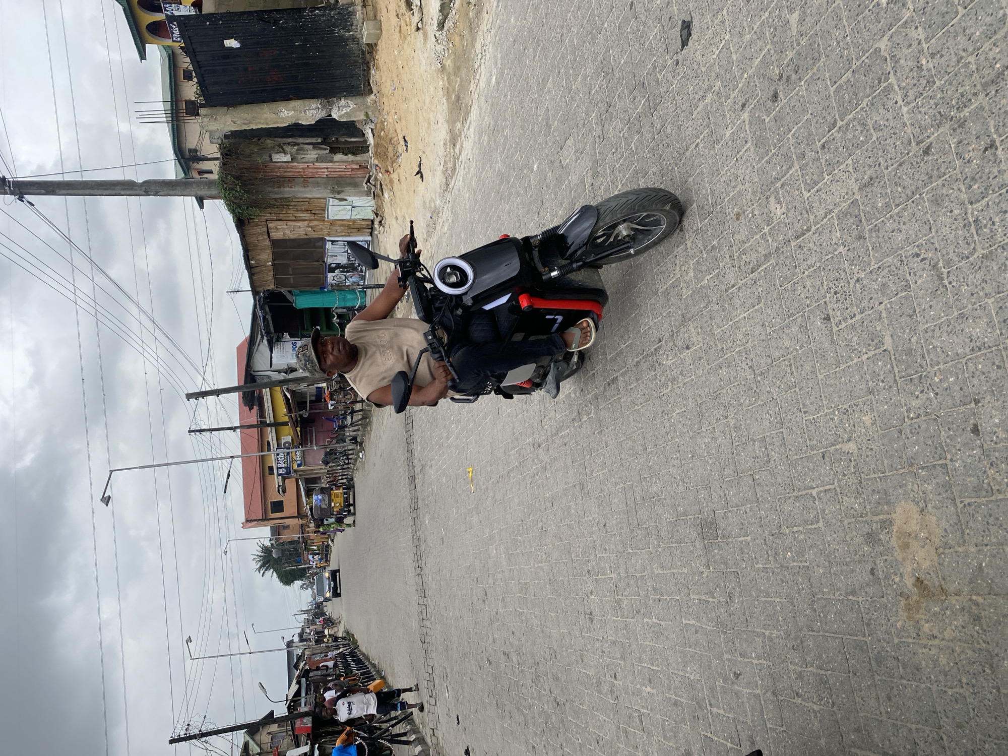 A man wearing a beige sleeveless shirt, dark pants, a camouflage baseball cap, and sandals is riding a black electric scooter down a paved street. The scooter has a circular white headlight with a ring of lights around it and red accents on the sides. The man is looking forward. The street is lined with buildings, some of which appear to be shops with metal fences and corrugated iron roofs. There are power lines overhead. In the background, a few other people and a yellow tricycle are visible, along with a dark-colored car driving away from the viewer. The sky is overcast. The scene appears to be a street in a moderately populated urban area, possibly a market street, with a mix of residential and commercial structures.
