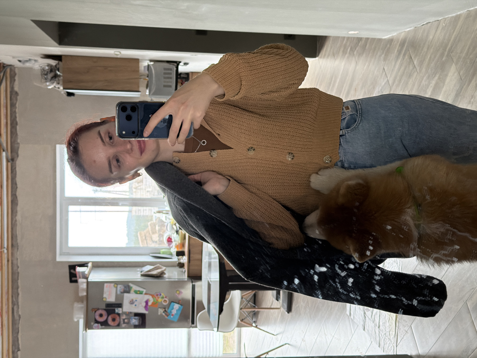 A young woman with reddish-brown hair is depicted taking a mirror selfie in what appears to be a home in Chaikovsky, Russia. She is wearing a light brown knitted cardigan with circular buttons and blue jeans, holding a dark smartphone, likely an iPhone, in her right hand to capture her reflection. A subtle smile plays on her lips, indicating a casual or content mood.
Beside her, a light brown dog, possibly a puppy, is partially visible, resting or sitting on a dark, possibly patterned, blanket or piece of fabric. The dog has distinct white markings or specks on its fur, particularly around its head and back, which might be paint or dust.
The background, visible through the mirror, suggests an indoor living space, likely a kitchen or dining area. A large window on the left allows natural daylight into the room, indicating it is daytime. Below the window, a stainless steel refrigerator is decorated with various colorful magnets, including one that appears to show two donuts. Adjacent to the fridge are kitchen counters and cabinets. To the far left of the image, exposed framing on a wall indicates ongoing renovation or construction. The floor is covered with light-colored rectangular tiles. No significant discernible text is clearly visible other than patterns on some fridge magnets and a small tag on the woman's jeans.