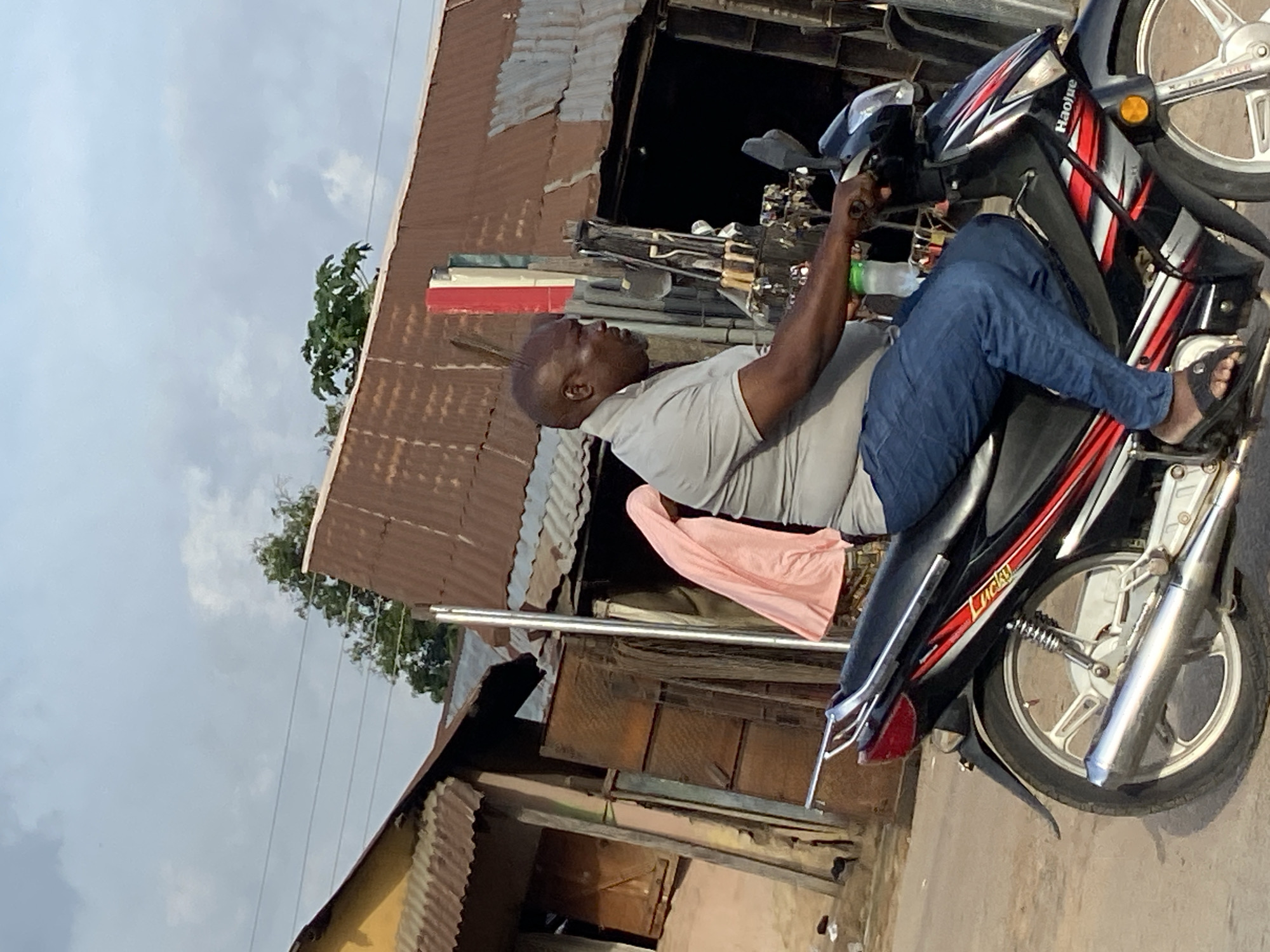 A dark-skinned man, appearing middle-aged and bald, is depicted seated on a motorcycle in an outdoor, urban setting. He wears a light gray t-shirt, blue jeans, and black open-toed sandals. He grips the handlebars of a black, red, and silver moped-style motorcycle. The brand name "Haojue" is visible on the front fairing, and "Lucky" is inscribed on a side panel near the rear. A green plastic water bottle is tucked into the motorcycle's front compartment.
The background features several structures with rusty, corrugated metal roofs and walls, suggesting older or informal buildings. A piece of pink fabric is draped or hanging behind the man. Green foliage is visible above the buildings and against the sky. Overhead power lines stretch across the pale, overcast sky, indicating it is daytime. The man's expression is neutral, and he appears to be either paused, waiting, or preparing to ride. The scene conveys a typical street view in a location such as Mararaba, Nigeria.