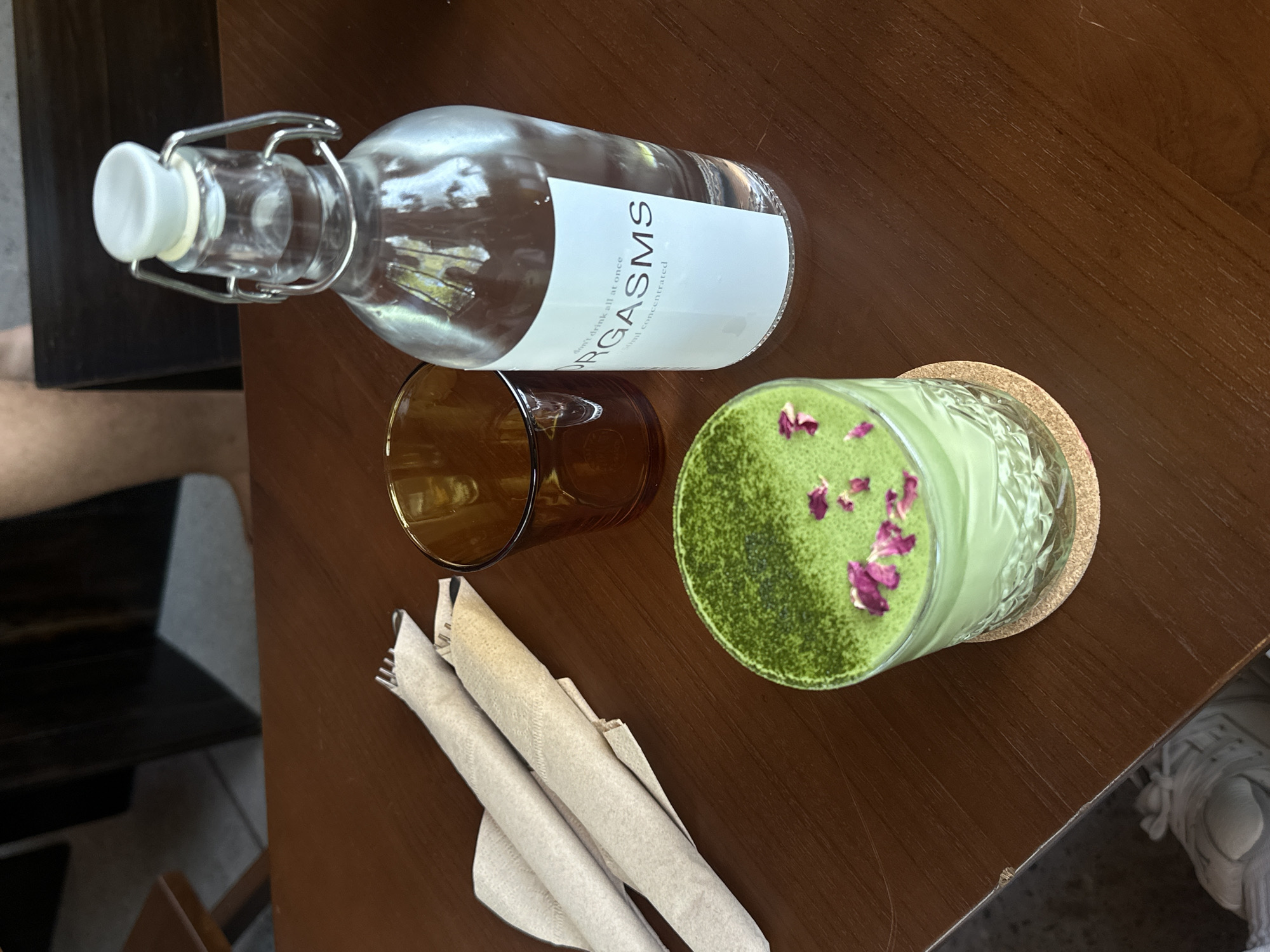 The image depicts a top-down view of a wooden table at an outdoor cafe in Unknown, Unknown. The table is set with a glass of green matcha, a brown glass, two wrapped utensils, and a clear glass bottle filled with water. The matcha is topped with a dusting of matcha powder and dried rose petals. The water bottle has a white label with text that reads "don't drink all at once" and "RGASMS" and "50ml concentrated". In the background, a person's legs and torso are visible, suggesting they are seated at the table. The cafe appears to have a casual, modern aesthetic.