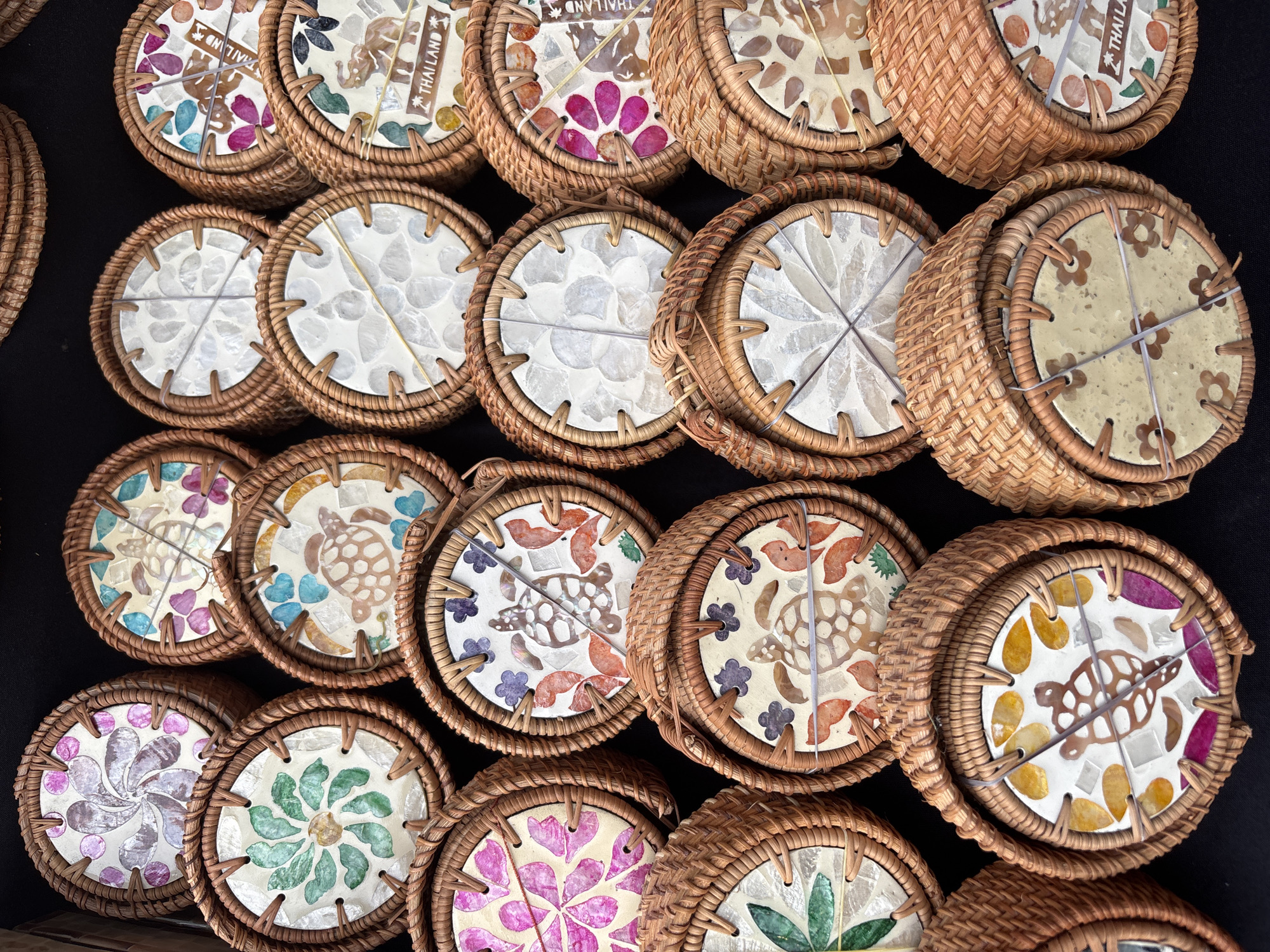The image shows a collection of round, woven baskets, likely coasters, displayed on a dark surface. Each coaster has a different mosaic design on the top, including floral patterns, turtles, and the word "Thailand" with an elephant image. The baskets are made of tightly woven brown material and are held together with thin rubber bands. The location is Baan Bo Phut, Thailand, as some coasters include that word.